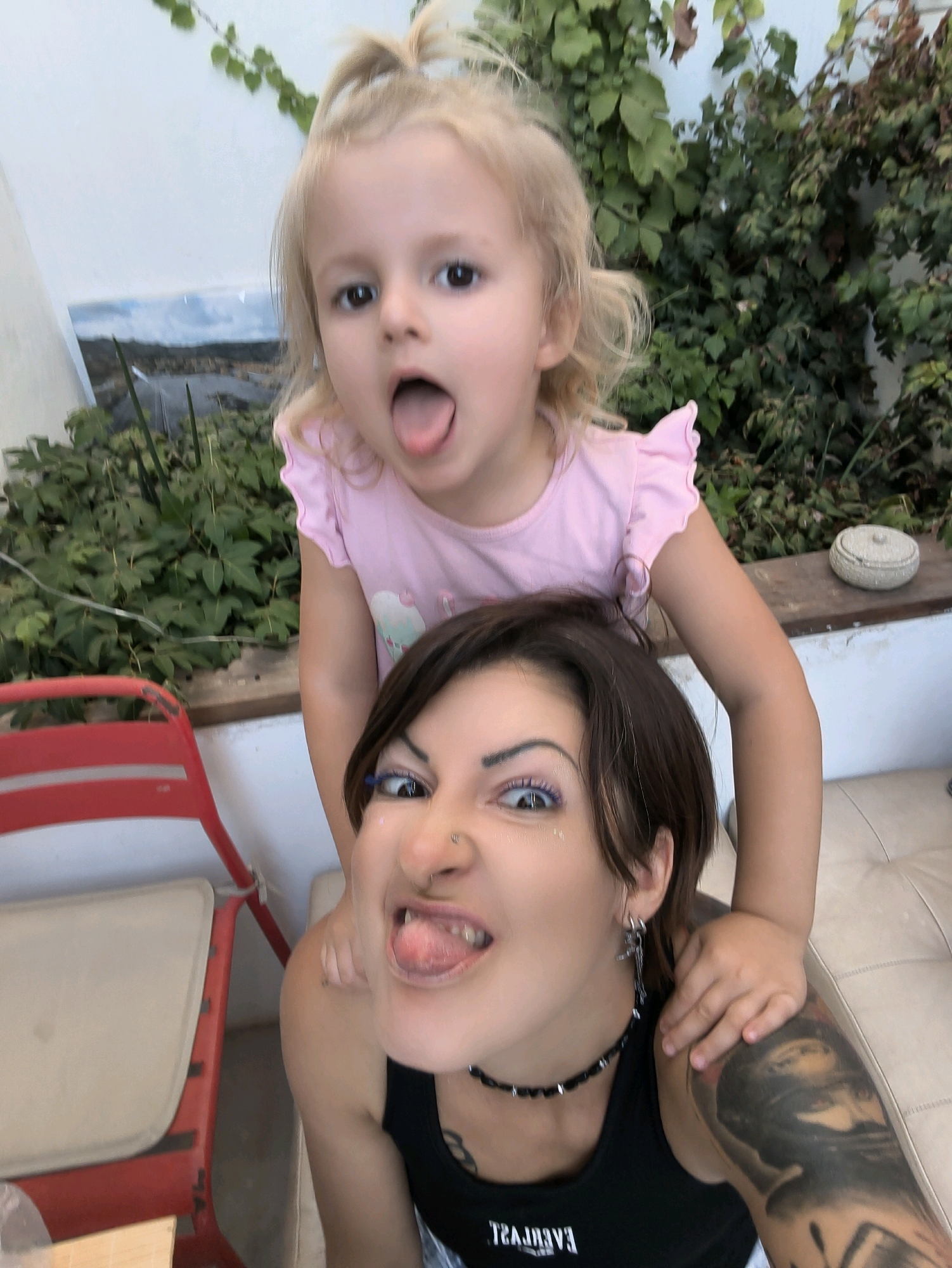 In an outdoor setting, a young child with blonde hair pulled into a ponytail and a pink t-shirt is on the shoulders of an adult. The child has their mouth open and tongue sticking out. The adult, with dark hair and a black tank top, is looking towards the camera with their mouth open and tongue sticking out. The adult has tattoos on their arm and is wearing a black choker necklace and earrings. Behind them is lush green foliage, a light-colored wall, and a red chair. A portion of a beige sofa is also visible. The scene appears to be a candid moment captured in a residential or garden area, potentially in or around Lachish Regional Council, Israel, given the implied geographic context.