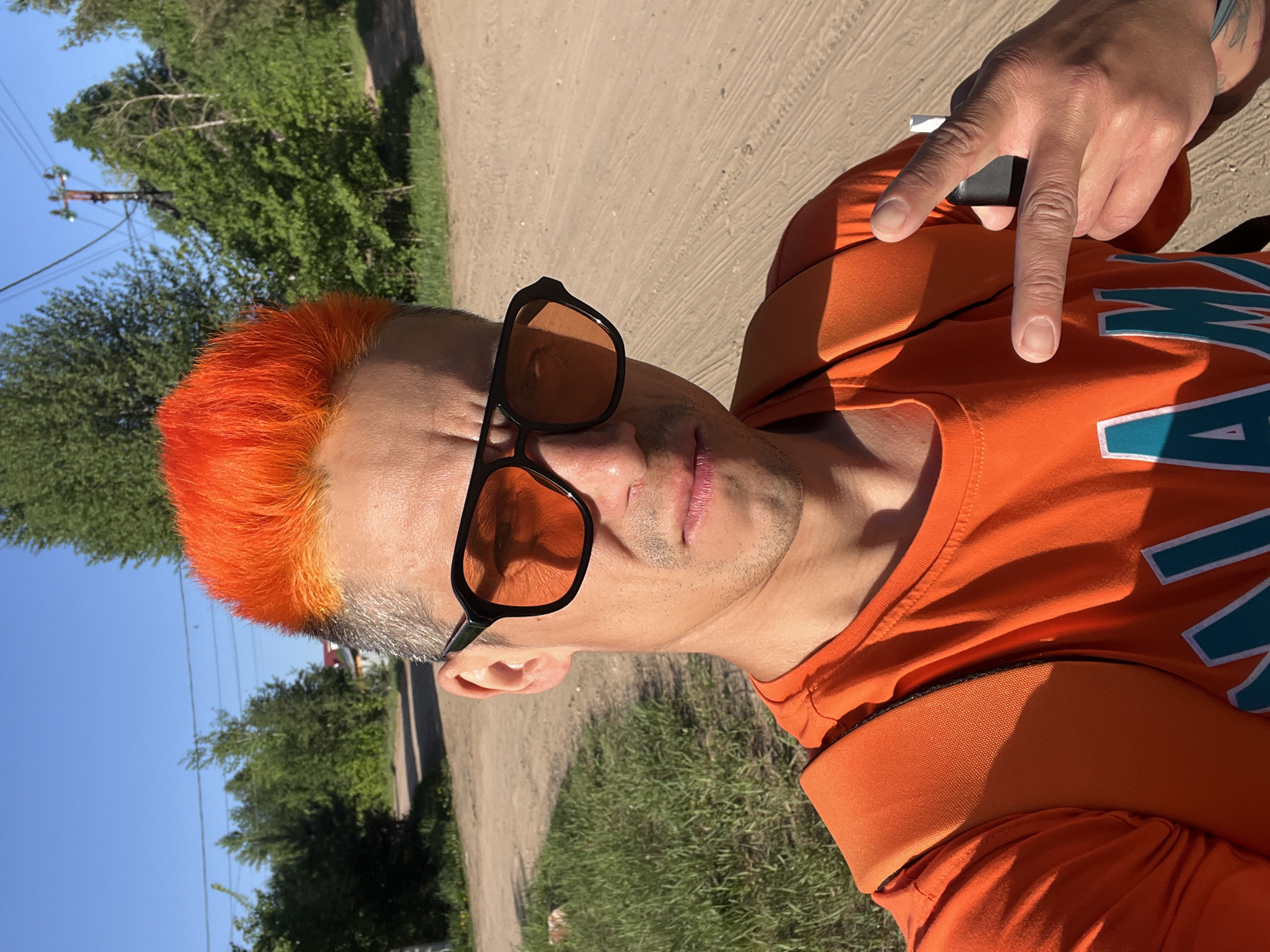 A man stands on a dirt road in an outdoor setting. He has orange hair, sunglasses, and an orange t-shirt. He is holding what appears to be a small, dark object between the fingers of his raised right hand. The environment includes trees, grass, a dirt road, and what appears to be power lines in the background. The setting is presumed to be in Chaikovsky, Russia, based on the user's prompt.