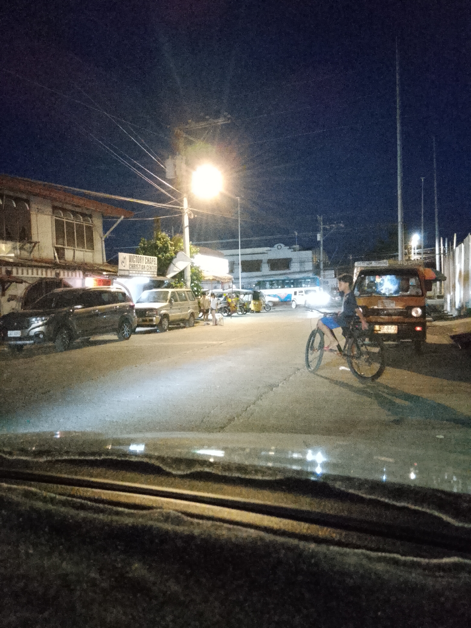 The image depicts a street scene at night in Davao City, Philippines. In the foreground, the hood of a vehicle is visible, with the interior slightly obscured. The street is paved and illuminated by streetlights and lights from vehicles and buildings.
On the left side of the street, a dark-colored SUV is parked, facing away from the viewer. To its right, a light-colored van or SUV is also parked. Further down the street, there is a building with a sign that reads "VICTORY CHAPEL CHRISTIAN CENTER". Several people are visible near the parked vehicles, some appearing to be talking.
In the middle ground, a young person is riding a bicycle, facing the camera and looking to their right. They are wearing a dark t-shirt and blue shorts. Behind the cyclist, a small orange van is parked, with its headlights on.
In the background, other vehicles are present, including what appears to be a bus and some tricycles. Streetlights and utility poles are scattered throughout the scene. The sky is dark, indicating nighttime. The overall impression is a typical urban street at night, with some activity and vehicles present.