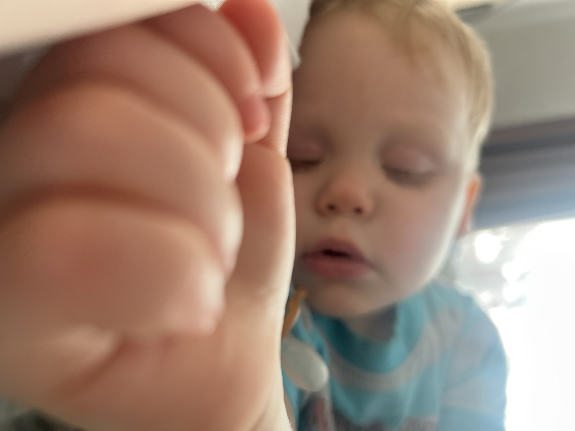 The image shows an infant or toddler, with light-colored hair, sleeping indoors in Chaikovsky, Russia. The child's head rests on white bedding, with eyes closed and mouth slightly ajar. A blue and white striped garment is visible on the child's upper body. A white, oval-shaped pacifier with an orange/brown strap is positioned near the child's mouth/chest. In the foreground, a child's partially clenched hand is prominently out of focus. The background is blurred and brightly illuminated, indicating an ambient light source.