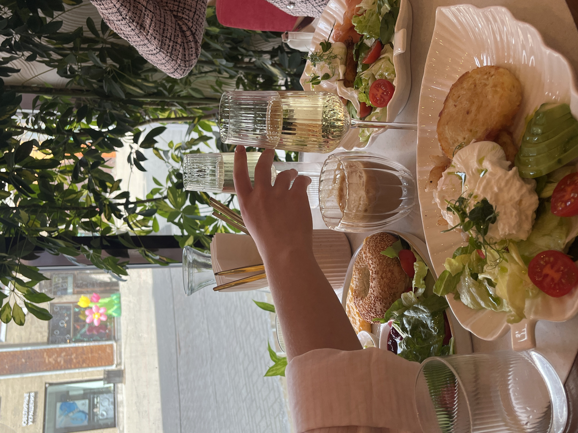 A partially visible person's arm, covered by a pale pink sleeve, reaches towards a glass of champagne. In the foreground, a plate holds a breakfast spread featuring a poached egg, shredded cheese, avocado, cherry tomatoes, and a piece of hash brown. Another plate in the background displays a sesame bagel, salad greens, and jam. Two flutes of champagne are visible, along with a glass of iced coffee. The setting appears to be a cafe, with large windows offering a view of a street scene. The exterior shot shows a building with a storefront and colorful flower decorations. Green plants are abundant inside the cafe, creating a vibrant atmosphere. The table is set with a white tablecloth.