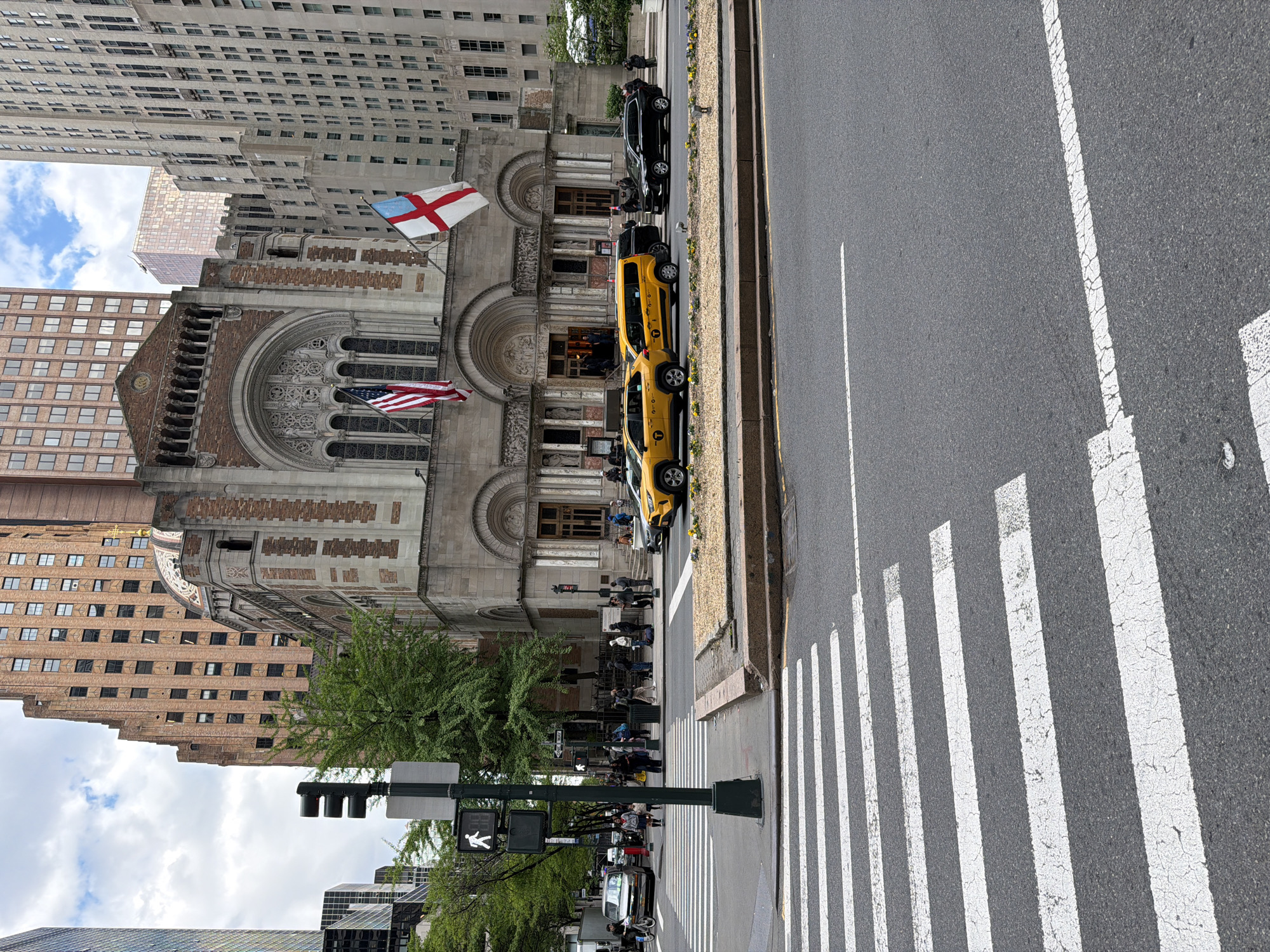 The image depicts a street scene in New York City, United States of America, specifically focusing on the Brick Presbyterian Church. The church is a prominent architectural feature with ornate detailing and two flags displayed: an American flag and another flag with a white background and red cross. The street shows typical urban activity. Pedestrians are gathered near the church entrance and along the sidewalk. Several yellow taxis and a black car are parked along the curb. A crosswalk is prominently visible in the foreground, marked with white stripes on the asphalt. Traffic lights and street signs are present, indicating a functioning urban environment. The background features tall buildings, typical of the New York City skyline. The sky is partly cloudy.