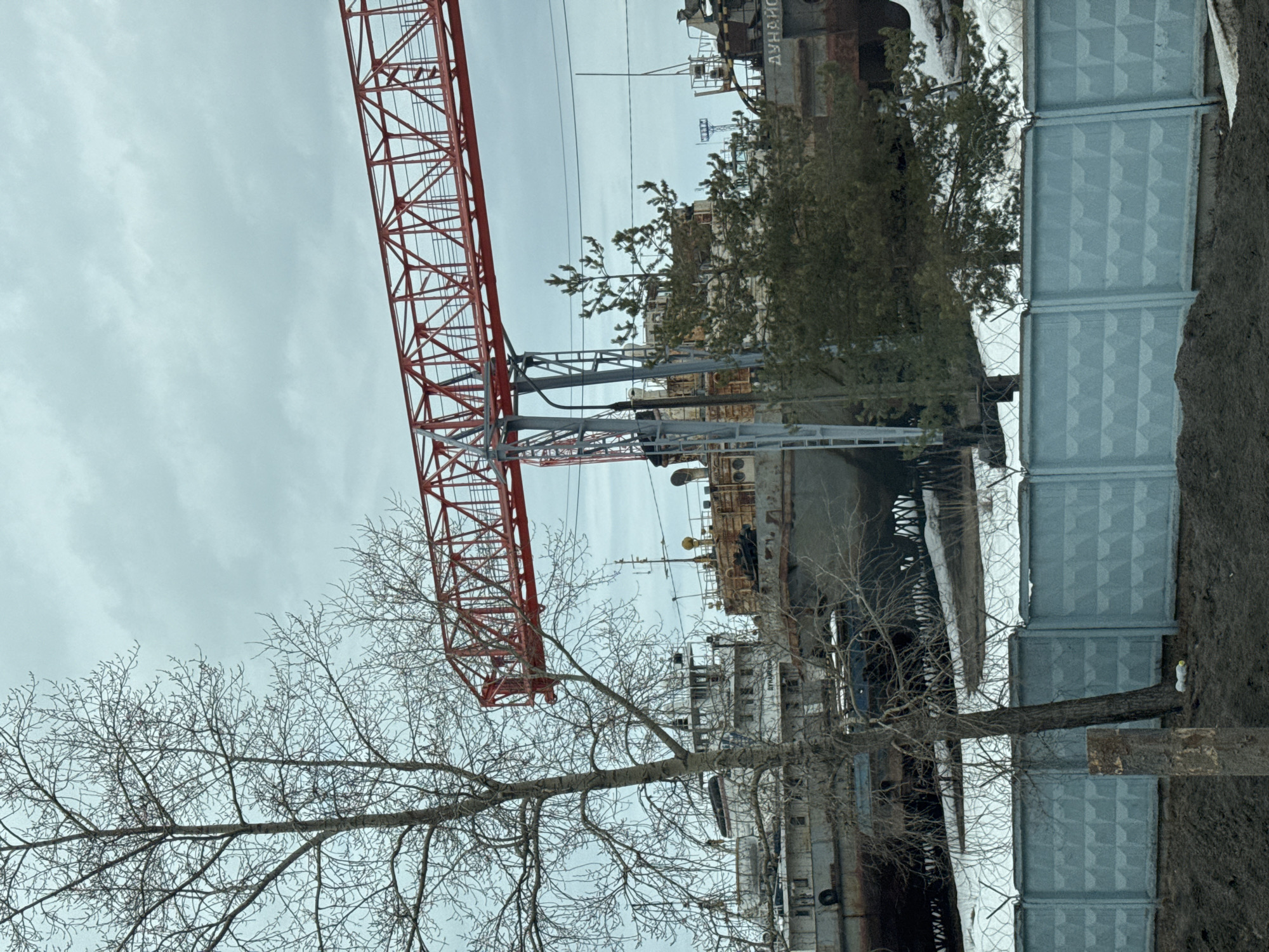 The image shows a view in Chaikovsky, Russia, capturing a scene with a red lattice crane extending diagonally across the upper left portion of the frame. Below the crane is industrial equipment behind a fence. Bare trees are prominent in the foreground and partially obscure the crane's base. The sky is overcast. The setting appears to be an industrial area or a shipyard. A concrete fence is visible on the right.