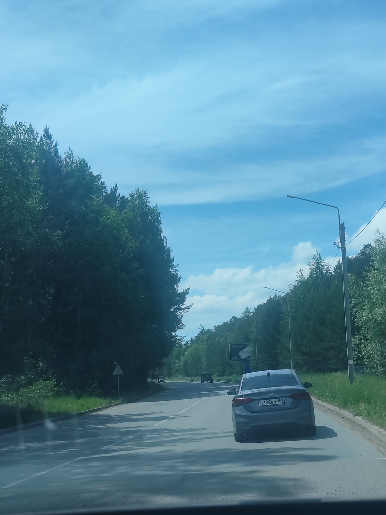 The image captures a daytime scene from inside a moving vehicle, looking forward down an asphalt road. A gray Hyundai sedan with Russian license plate "H192BX 159" is prominently featured in the foreground, moving away from the viewer. The road, which curves gently to the left in the distance, is lined on both sides by dense green trees, suggesting a forest or wooded area. Sunlight casts distinct shadows of trees and the car onto the road surface.
The sky above is bright blue with scattered white clouds. Several streetlights on tall poles are visible along the right side of the road, and another appears further down on the left. A triangular warning road sign is present on the left side of the road amidst the foliage. In the distance, on the right, a billboard can be seen, though its text is mostly illegible, with only what appears to be "101" and some Cyrillic characters vaguely discernible. Another dark-colored vehicle is visible further down the road on the left. The scene depicts travel through a well-maintained road in a natural setting, consistent with the given location context of Chaikovsky, Russia, as indicated by the Perm Krai region code "159" on the license plate.