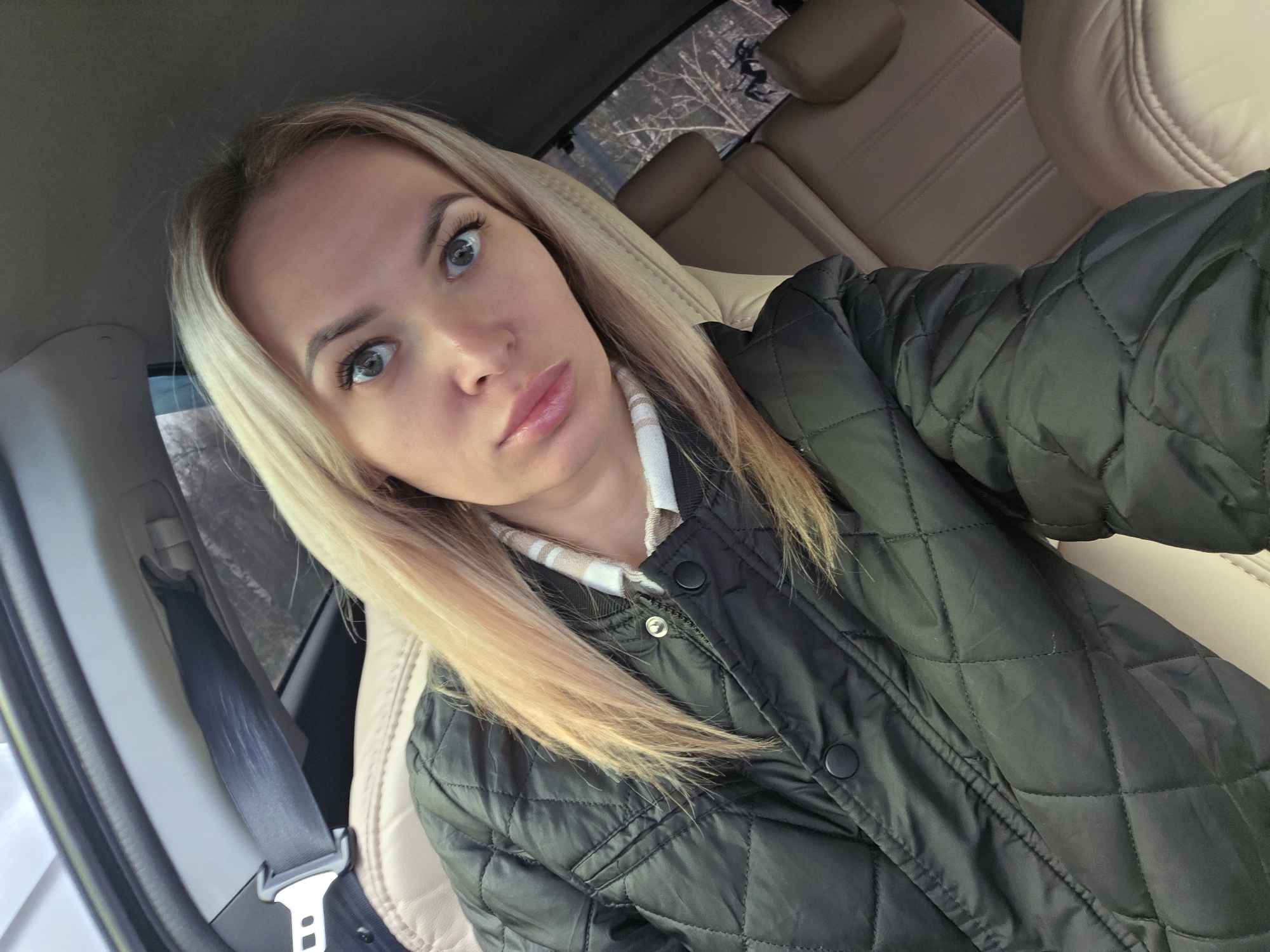 A young woman with blonde hair and blue eyes is taking a selfie in the passenger seat of a car. She is wearing a black quilted jacket and a collared shirt with a light-colored pattern. The car has beige leather seats, and the seatbelt is visible. Outside the car window, bare trees can be seen, suggesting it is winter or early spring. The city of Chaikovsky, Russia, is not discernible from the visual information provided.