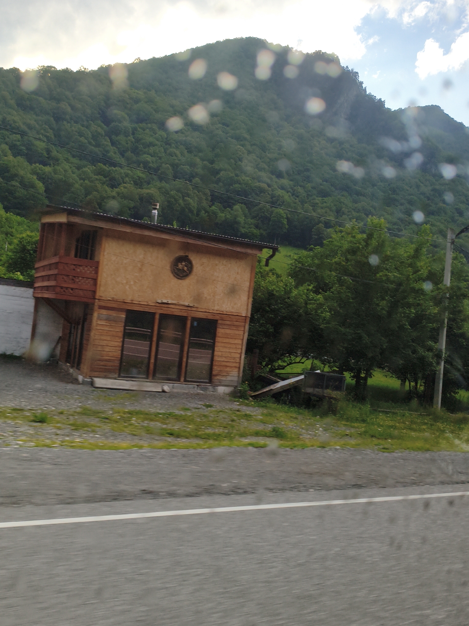 A wooden building with a balcony and large windows sits on a gravel lot next to a paved road in Gusyra, Russia. The building is situated at the base of a steep, tree-covered mountain. The sky is cloudy with patches of blue.