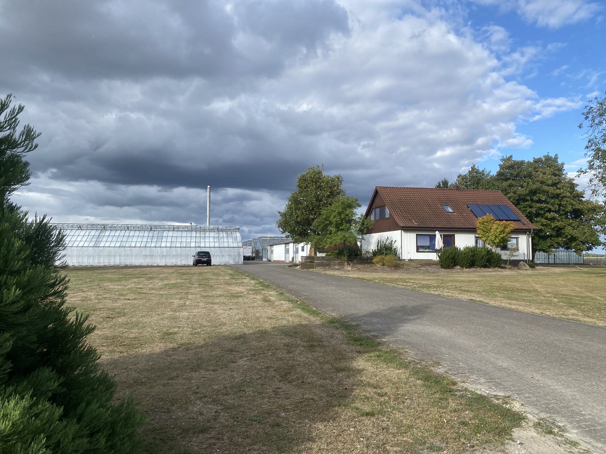 In Schwarme, Germany, a suburban or rural scene unfolds under a dramatic sky. A vast greenhouse structure, made of glass and metal, dominates the left side of the image, with a single dark car parked in front of it. To the right, a detached house with a red-tiled roof and solar panels sits on a manicured lawn, bordered by trees. A paved driveway leads from the foreground to the house, dividing the property. The sky above is a mix of dark, heavy clouds and patches of bright blue, suggesting a dynamic weather system. The overall impression is one of a functional, residential property with agricultural or industrial elements, set against a backdrop of impressive cloud formations.