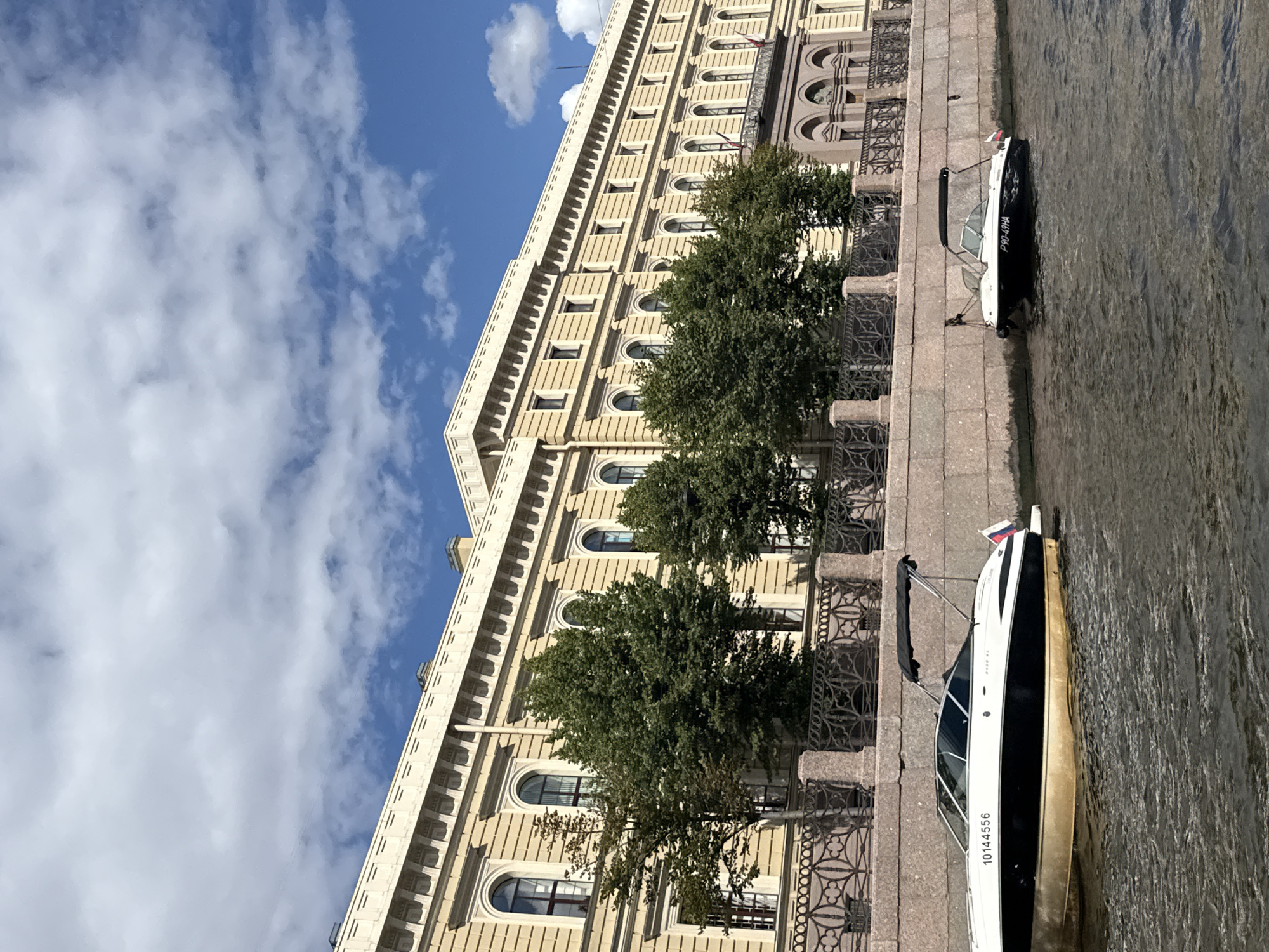 This image depicts a canal in Saint Petersburg, Russia, with a large, pale yellow building lining the opposite bank. The building is multi-story, with numerous arched windows and decorative architectural elements. Trees are visible in front of the building, partially obscuring its lower floors. A stone embankment runs along the canal, topped with an ornate metal fence.
Two speedboats are present in the canal. The first, on the left, is white and black with a gold hull. It has a registration number "10144556" visible on its side and a Russian flag at its stern. The second speedboat, on the right, is also white and black, with a black canopy. It has a registration number "P90-49HA" visible on its hull and a Russian flag at its stern.
The water in the canal is dark and rippled, reflecting the light from the sky. The sky above is blue with scattered white clouds. The overall scene suggests a sunny day, likely during the warmer months in Saint Petersburg.