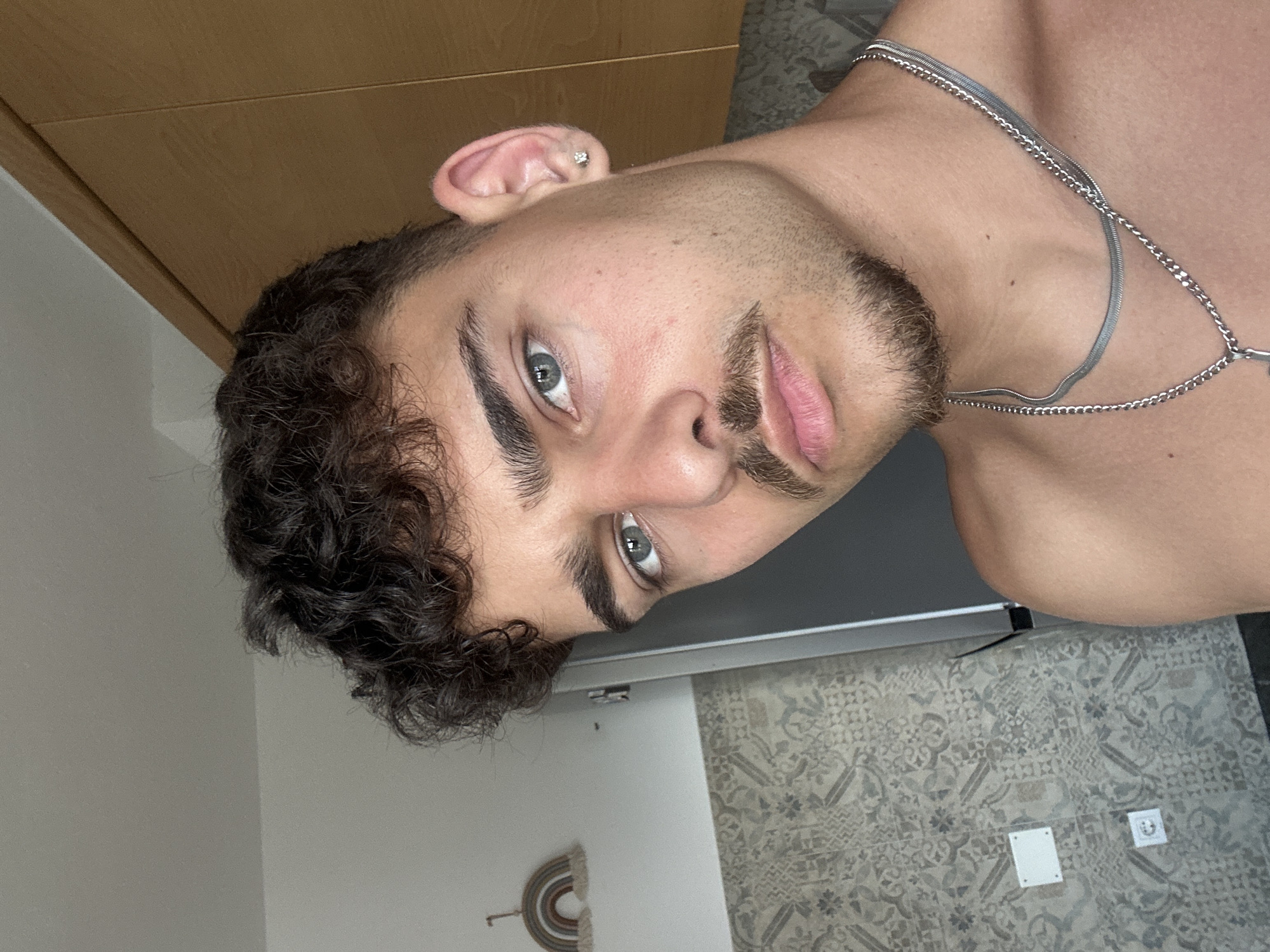 A close-up, angled selfie of a light-skinned male with dark curly hair, a neatly trimmed beard, and light-colored eyes. He is wearing a silver chain necklace. The background includes a wooden cabinet, a white wall with a decorative rainbow hanging, and patterned grey flooring. No evidence indicates the image was taken in Troyan, Bulgaria.