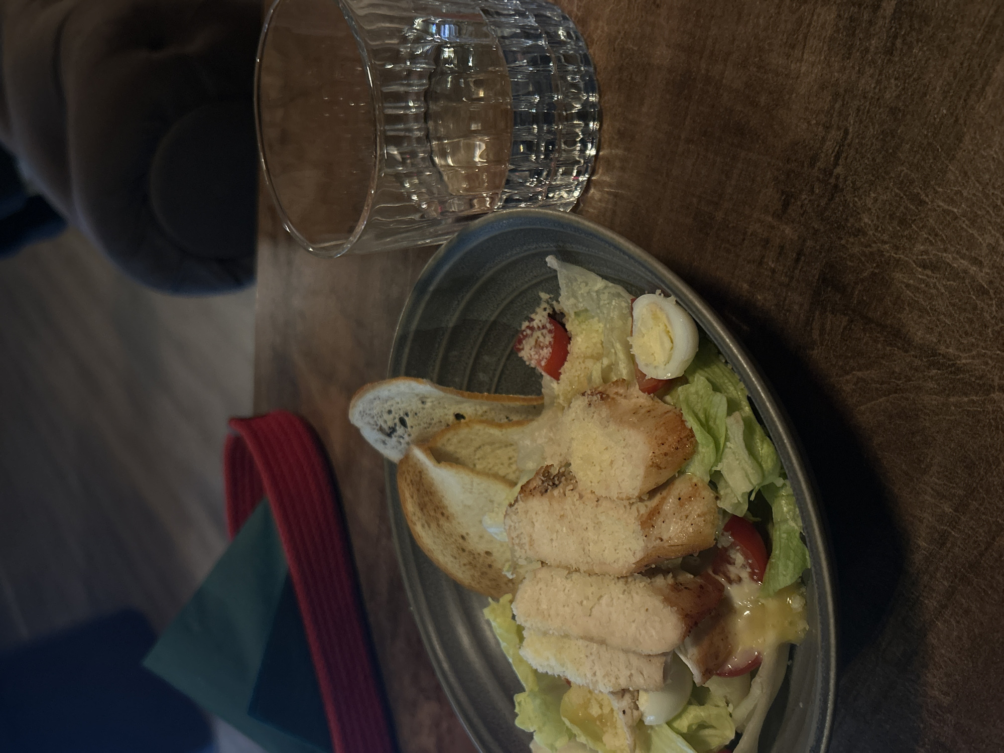 The image shows a Caesar salad served on a gray plate, positioned on a wooden table next to a textured glass of water. The salad consists of lettuce, sliced grilled chicken, croutons, halved eggs, tomatoes, and what appears to be Parmesan cheese. A red napkin is visible near the bottom left corner. The background includes a glimpse of a person's shoulder. The location is not discernible from the image.