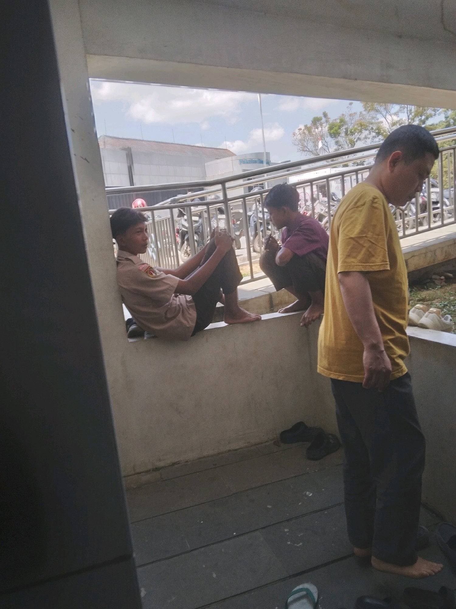 In the city of Sokaraja, Indonesia, three males are visible in an outdoor, covered area. The individual on the left, a young male wearing a light brown uniform shirt with an emblem, is sitting on a raised concrete ledge, leaning against a dark gray column, with his legs extended and crossed at the ankles. To his right, a second young male, wearing a purple t-shirt and dark pants, is squatting on the same ledge. On the far right, an adult male in a yellow t-shirt and dark pants stands facing away from the viewer, his right arm resting on his hip. The background reveals a metal railing, parked motorcycles, trees, and buildings under a cloudy sky. The ground in the foreground is a wooden planked surface with several pairs of footwear, including dark sandals and light-colored clogs, scattered around.