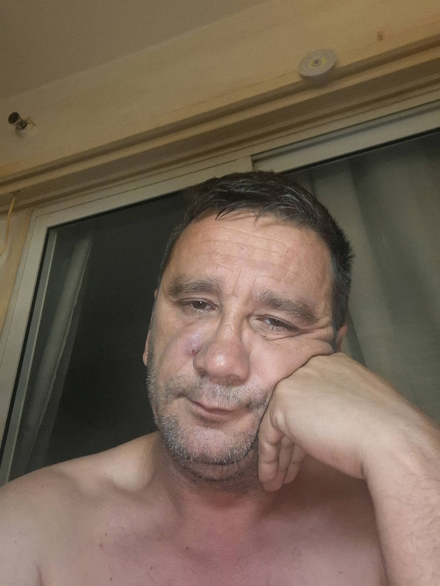 A bare-chested adult male, facing forward and resting his right hand on his cheek, is captured in a close-up, high-angle, indoor shot. The man, who appears to be in his 40s or 50s, has short, dark hair with some graying, stubble on his chin, and a pensive expression. His right eye is slightly squinted. The background consists of a window with vertical blinds or a door with a dark curtain, and above, a cream-colored ceiling with molding and a light fixture. The overall lighting suggests an indoor environment. The image does not contain information that allows for specific geographic localization within Yerocham, Israel.