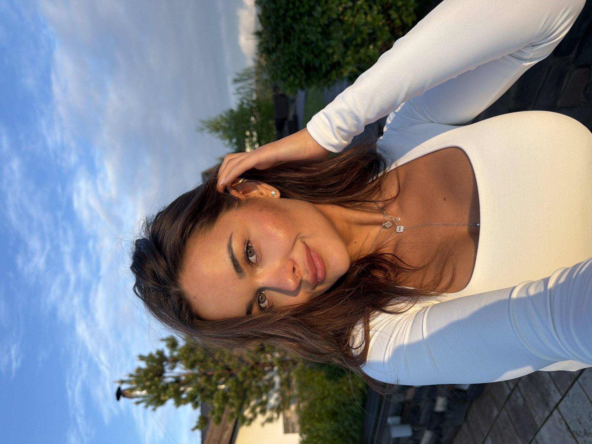 A young woman poses outdoors in Элита, Russia. She wears a white, long-sleeved top and a delicate silver necklace. Her brown hair is styled loosely, and she touches it with her right hand. The background features a clear blue sky with some clouds, trees, and building rooftops. The ground appears to be brick or stone.