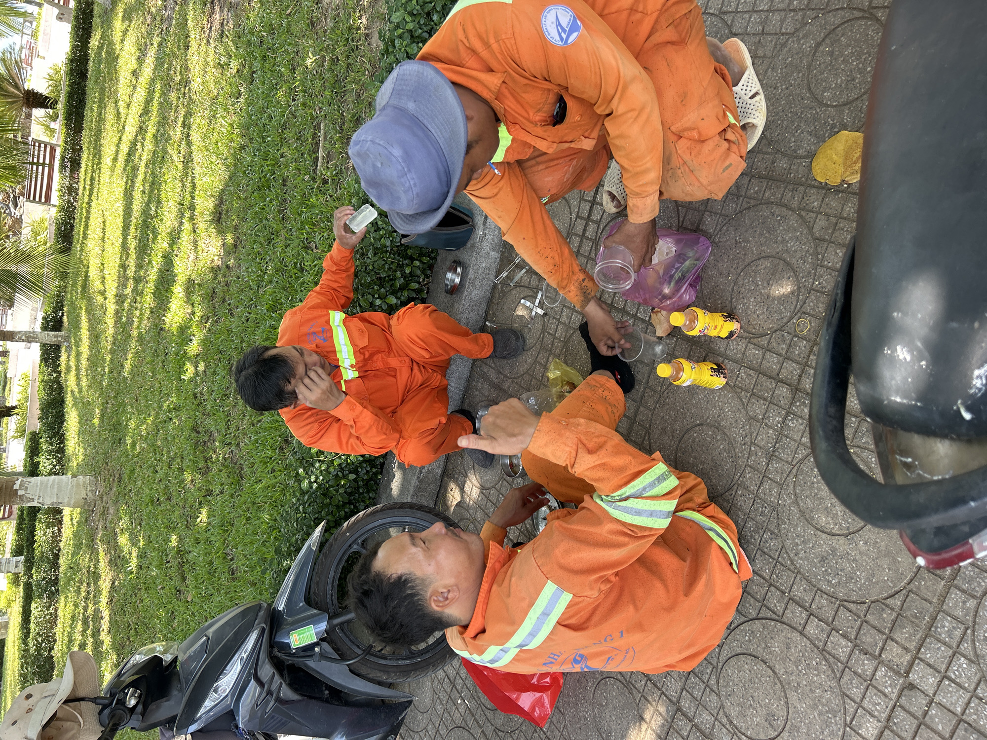 Three men in bright orange work jumpsuits with reflective green and grey stripes are taking a break on a tiled sidewalk next to a grassy area with palm trees and bushes. The scene is illuminated by bright sunlight, casting distinct shadows on the grass, suggesting it is midday or early afternoon. The setting is an urban park or public space in Nha Trang, Vietnam.
One man, wearing a blue bucket hat, is sitting with his back partially to the camera, reaching into a purple plastic bag on the ground. He has a clear plastic cup in his hand. Near him are several small yellow bottles, likely beverages, and other clear plastic cups. Another man is sitting opposite him, holding a piece of ice or a clear ice block to his face, appearing to be resting or cooling down. The third man is lying on his side in the foreground, looking up, with his arm bent.
A black scooter or motorcycle is partially visible in the left foreground, with a light-colored hat resting on it. The men's orange uniforms show some dirt and stains, indicating recent work. The activity appears to be a refreshment break from their duties, possibly related to municipal or park maintenance.
Visible text includes a circular logo on the orange jumpsuit of the man with the blue hat, which contains Vietnamese text such as "CÔNG TY CỔ PHẦN" (Joint Stock Company) and "CÔNG TRÌNH CÔNG CỘNG" (Public Works). On the uniform of the man lying down, part of a logo that reads "NHA TRANG 1" is discernible, likely indicating their work area or team. Text on the yellow bottles and a green sticker on the scooter are present but not legible.