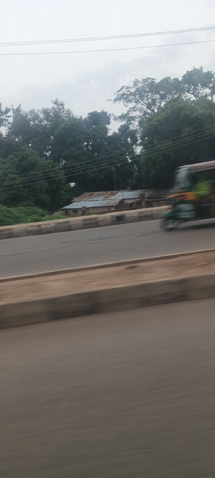 This image presents a candid roadside view in Jos, Nigeria, captured from a moving vehicle, as indicated by the noticeable motion blur across the road and a moving three-wheeled auto-rickshaw (likely a Keke NAPEP). The weather appears overcast, with a uniformly grey sky and no discernible shadows, suggesting a cloudy day.
The foreground is dominated by a multi-lane road, separated from the roadside by a low, light-colored concrete barrier. Beyond this barrier, the midground features several modest structures with corrugated metal roofs, some rusted and some newer, nestled amidst a dense expanse of green trees that form the background. These buildings appear to be simple dwellings or sheds. Two thin power lines are visible stretching across the upper mid-section of the image, horizontally.
On the right side of the road, a blurred auto-rickshaw, identifiable by its distinct shape and splashes of yellow and green color, is in motion. The activity depicted is everyday road travel, capturing a fleeting moment of the local environment. No distinct individuals or specific text are legible in the image. The scene portrays a functional, lived-in landscape typical of a semi-urban or rural area in the specified region.