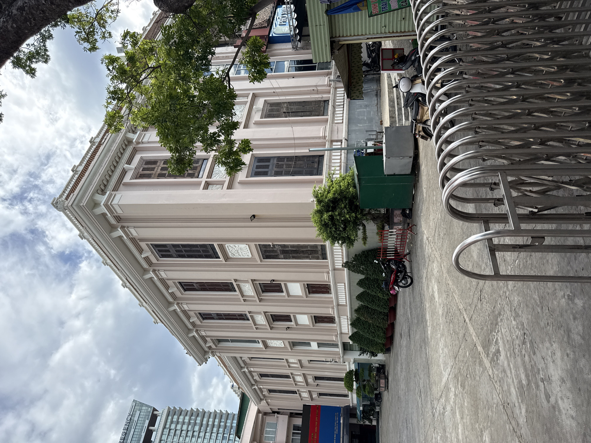 The image shows the facade of a large, light pink building with classical architectural elements. The building has multiple floors and numerous tall, narrow windows with dark shutters. Decorative moldings and pilasters adorn the exterior. In the foreground, a paved courtyard area is visible, with several potted plants and trees lining the building's base. A red motorcycle and other vehicles are parked in the courtyard. A silver retractable gate is prominent in the lower right corner of the image. In the background, a modern high-rise building is visible to the left. The sky is cloudy. The location is the Nha Trang Post Office building, located at 131 Thống Nhất Street, Nha Trang City, Khánh Hòa Province, Vietnam.