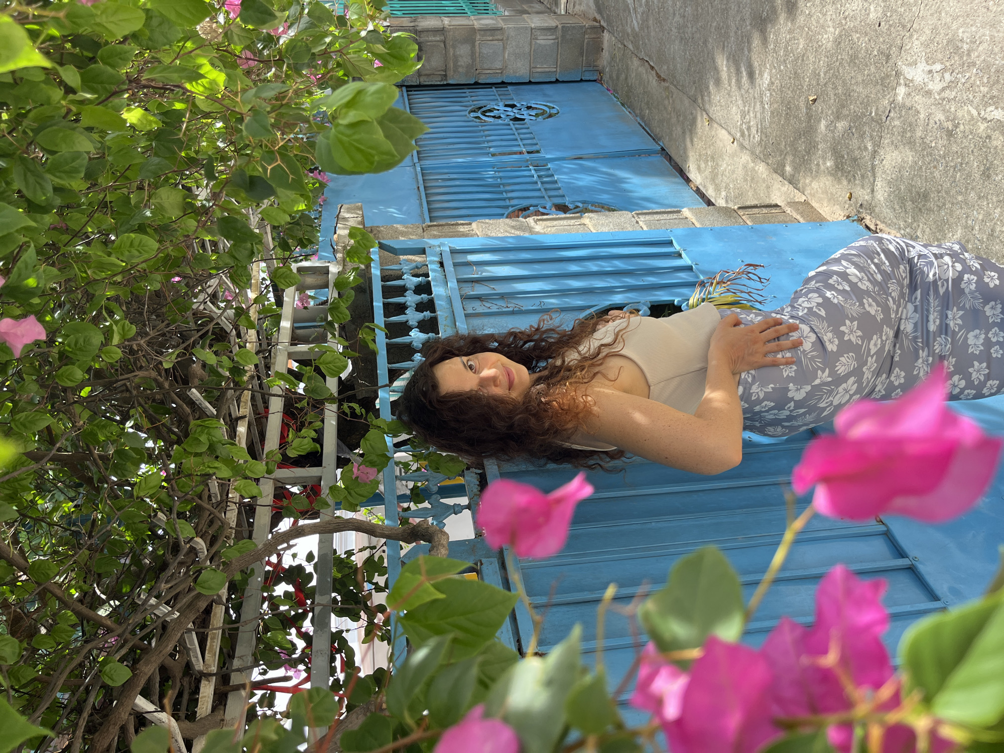 A woman with curly brown hair stands outdoors in Mũi Né, Vietnam, leaning against a blue metal gate. She is wearing a beige tank top and a light grey skirt with a white floral pattern. The gate is part of a stone wall, and behind it, a building is partially visible. Pink bougainvillea flowers and green leaves hang from a tree overhead, casting shadows on the scene. The ground is a concrete pathway.