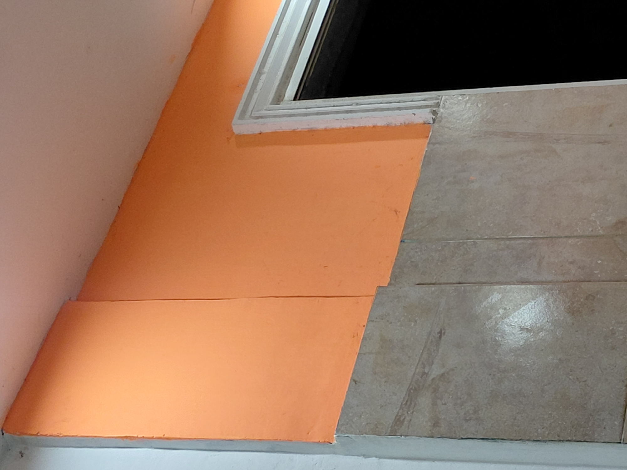 The image depicts an interior or semi-enclosed corner of a building in Santa Ana, Venezuela, focusing on architectural details.
**Subjects:** The primary subjects are a plain white wall on the left, an orange-painted wall, a white-framed window, and a section of light brown tiled surface. A thin, light-colored trim piece is visible at the very bottom edge of the frame. No people or animals are present.
**Activity or event taking place:** The image is a static shot of structural and decorative elements; no activity or event is occurring.
**Notable details:**
The white wall on the left has a slightly textured finish. It meets a vibrantly colored orange wall at an angle. The orange wall displays a distinct horizontal seam or line, suggesting two separate sections or a specific design choice. The paint finish on the orange wall shows some visible texture.
A window, framed with simple white molding, is set into the upper part of the orange wall. The opening of the window is completely black, indicating a dark interior space beyond, or that the image was taken at night.
To the right of the window, and continuing downwards, is a surface covered with rectangular, light brown or beige tiles. These tiles have visible grout lines and a somewhat glossy or textured finish, showing variations in tone and some minor wear.
A narrow, light-colored (possibly white or light blue) trim runs horizontally along the bottom of the image, separating the orange and tiled surfaces from a lower wall or base. The lighting is relatively even, without strong shadows, suggesting diffused light or indoor lighting.
**Visible text:** There is no visible text in the image.