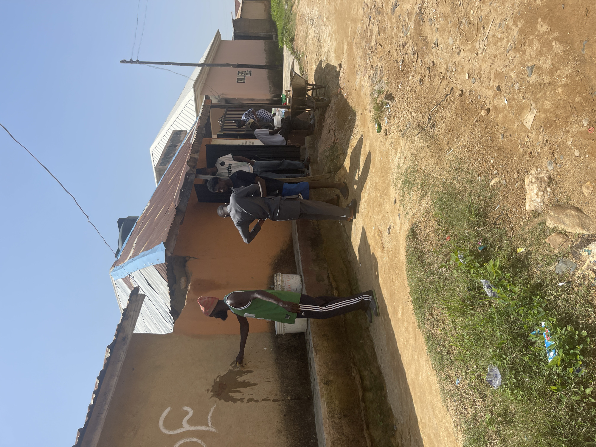 The image depicts an outdoor scene in what appears to be a village or informal settlement in Jos, Nigeria. Several people are present, with a focus on a group gathered near the entrance of a building.
The building has an orange-colored facade and a corrugated metal roof. Above the entrance, text is visible on the building, reading "CHICLES COLLEGE". A wheelbarrow is parked to the right of the entrance.
In the foreground, a man wearing a green basketball jersey, black track pants with white stripes, and a red beanie is leaning down to the ground, seemingly interacting with the wall or ground. Another person, dressed in a grey outfit, stands behind him, holding a stick or pole. Other individuals are standing or sitting near the building's entrance, some appearing to be in conversation.
The ground is a mixture of dirt, sand, and some patches of grass. Some litter, including plastic bottles, is visible on the ground and in the grass. The sky is clear and bright, suggesting it is daytime, likely a sunny day. The overall atmosphere appears casual, with people engaged in everyday activities. The architecture and the dry, dusty environment are consistent with a location in Nigeria.
