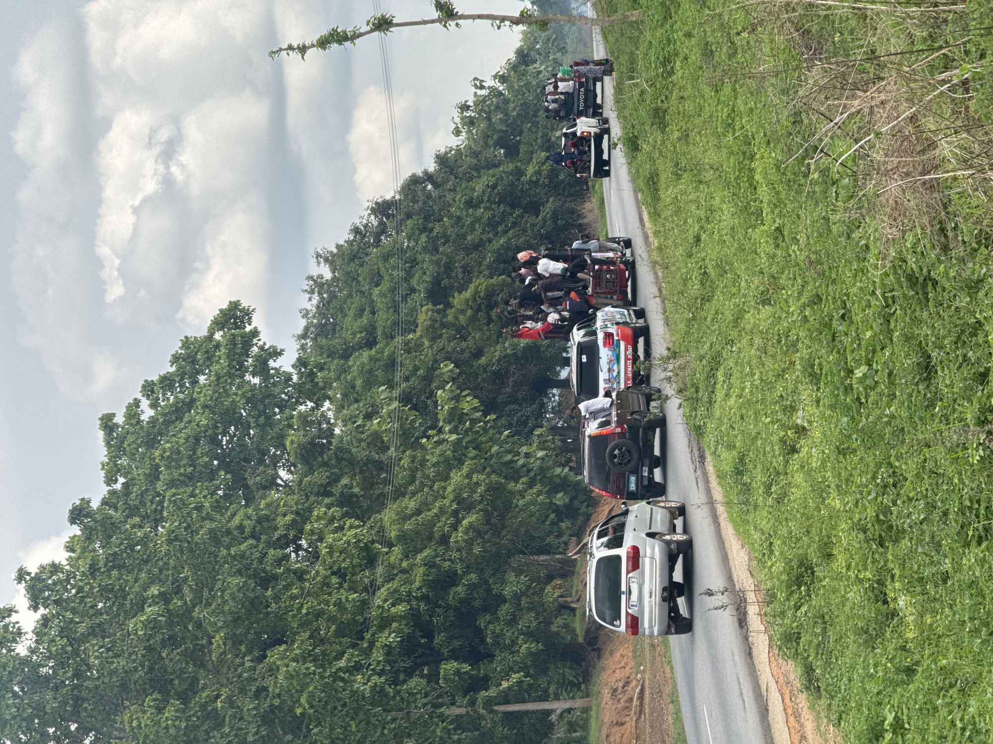 The image shows a procession of vehicles traveling along a road lined with trees and vegetation, likely on the outskirts of Akwanga, Nigeria. The vehicles include cars, pickup trucks, and auto rickshaws (tricycles), several of which are heavily laden with people. The sky is overcast. The location is identifiable as being near Akwanga in Nigeria based on unique auto rickshaw models in the parade.