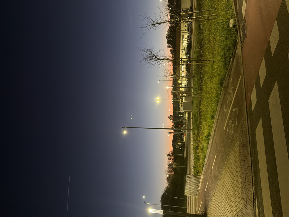 The image depicts an urban intersection in Penafiel, Portugal, at dusk or early night. The sky transitions from a deep navy blue at the top, featuring a faint white contrail in the upper left quadrant, to an orange-pink gradient above the horizon line. Two small, distinct point lights are visible in the upper-mid sky, to the right of a prominent streetlight.
In the foreground, a red-colored crosswalk with white stripes is present. To its immediate left, a textured, light-colored concrete walkway runs alongside a red-paved bicycle lane, marked with a white bicycle symbol and a dashed white line. A low metal barrier separates these paths from a central grassy median.
The midground features this grassy median containing several young, bare deciduous trees and multiple tall streetlights emitting bright yellow-white light. Beyond the median, a multi-lane road system is visible, populated by numerous parked vehicles and what appear to be traffic signals.
In the background, a large, single-story commercial building with a light-colored facade and a dark roof is illuminated, with a parking lot in front filled with various models of passenger cars. Distant hills covered in dark foliage are visible on the left side of the frame. No individuals are visible within the scene.