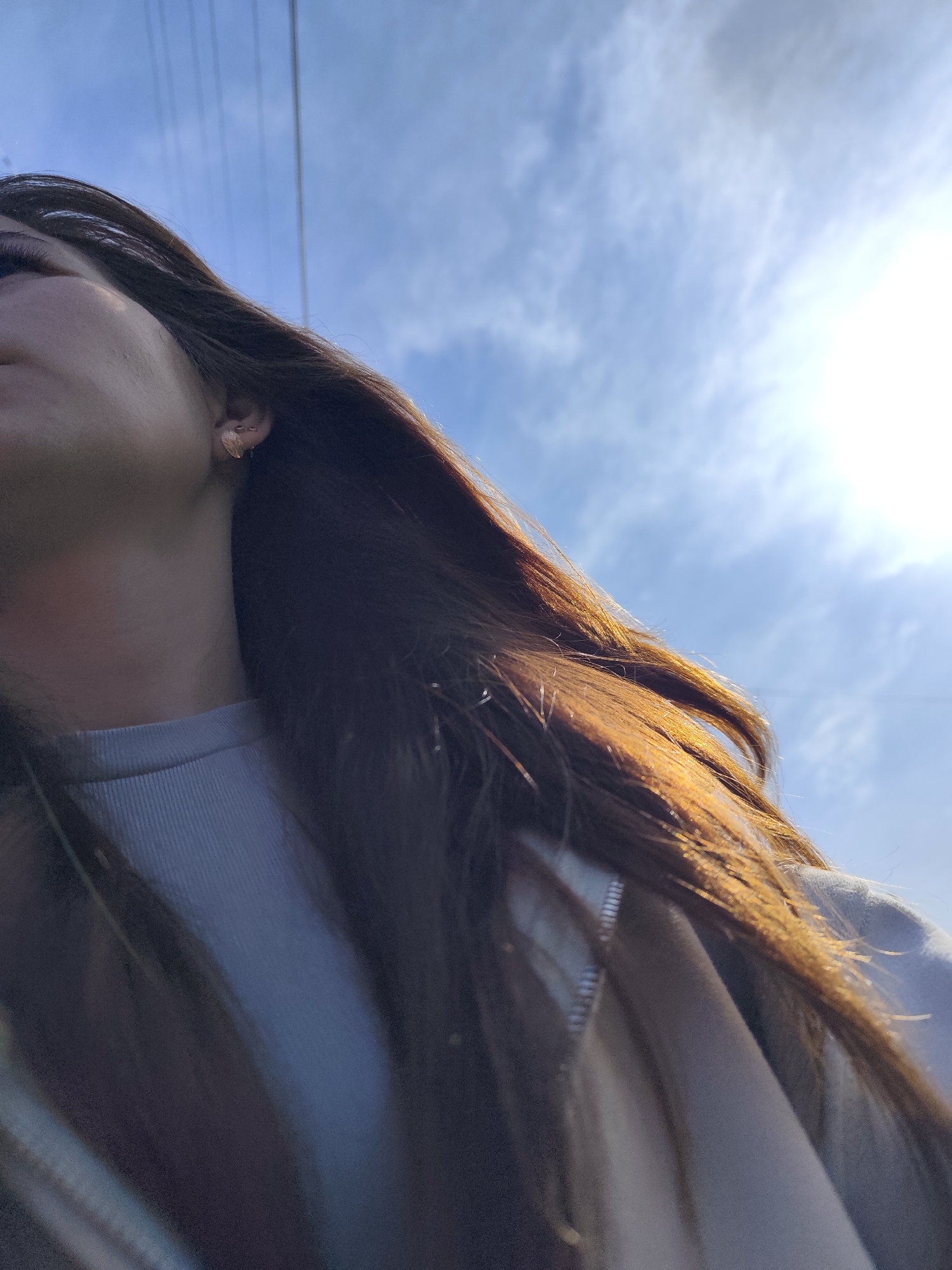 A low-angle shot shows a person with long brown hair, wearing a light-colored shirt or jacket, standing outdoors on a sunny day. The sky is visible with scattered clouds. Power lines stretch across the upper portion of the frame. No identifiable location. No action or interaction.