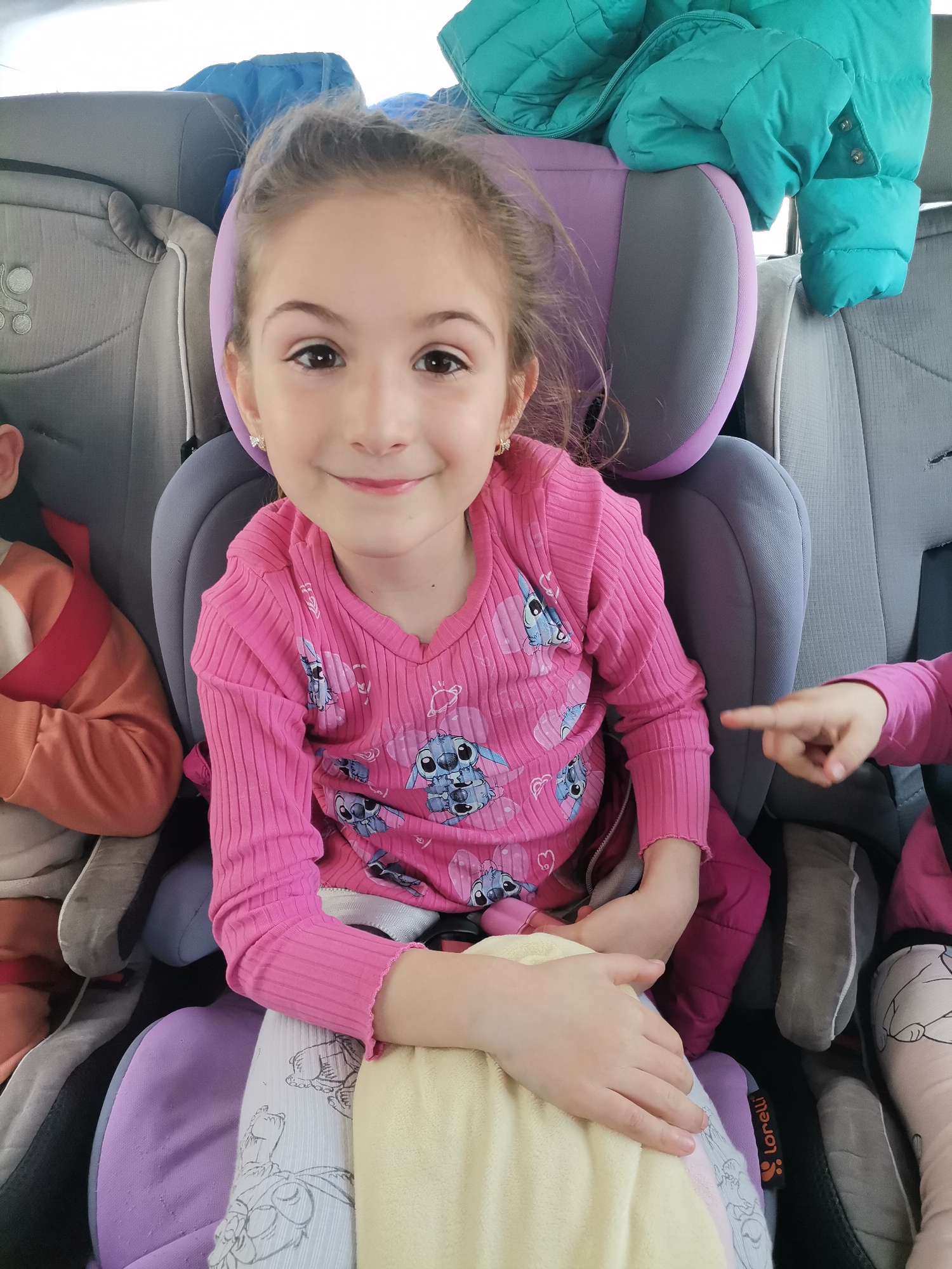 A young girl with brown hair pulled back in a bun sits in a gray and purple Lorelli brand car seat. She wears a pink long-sleeved shirt with a "Lilo and Stitch" cartoon pattern and white pants with cartoon sketches. The girl is smiling and looking directly at the camera. She wears small, decorative earrings. Another child is seated in a car seat to her left, visible by their red and orange sleeve. To the girl's right, another child is visible by their pointing finger and pink long-sleeved shirt. A blue and green jacket hangs over the car seat behind her. The environment appears to be the interior of a car. The location, Dobrich, Bulgaria, is not evident within the provided image.