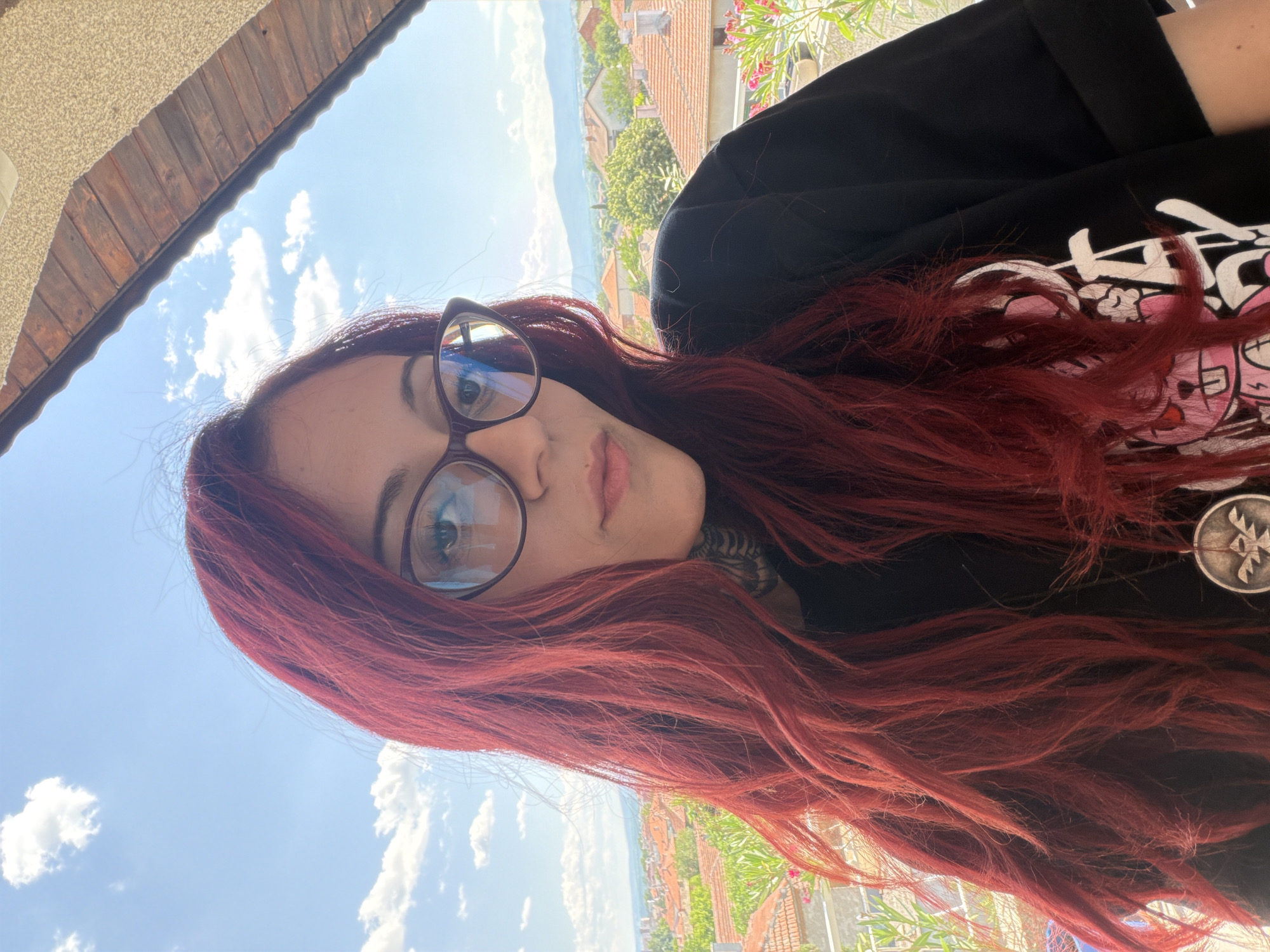 A woman with long, reddish-brown hair and glasses is taking a selfie outdoors. She is wearing a black t-shirt with a graphic design on it. The background shows a clear blue sky with some white clouds, and beyond that, a view of buildings with red-tiled roofs, trees, and a body of water in the distance. The scene appears to be set in a residential area, possibly on a balcony or patio, given the presence of a wooden overhang and some greenery. The lighting suggests it is daytime, likely sunny. The woman's expression is neutral, and the overall mood is casual. A silver circular object with a symbol is visible on her t-shirt.