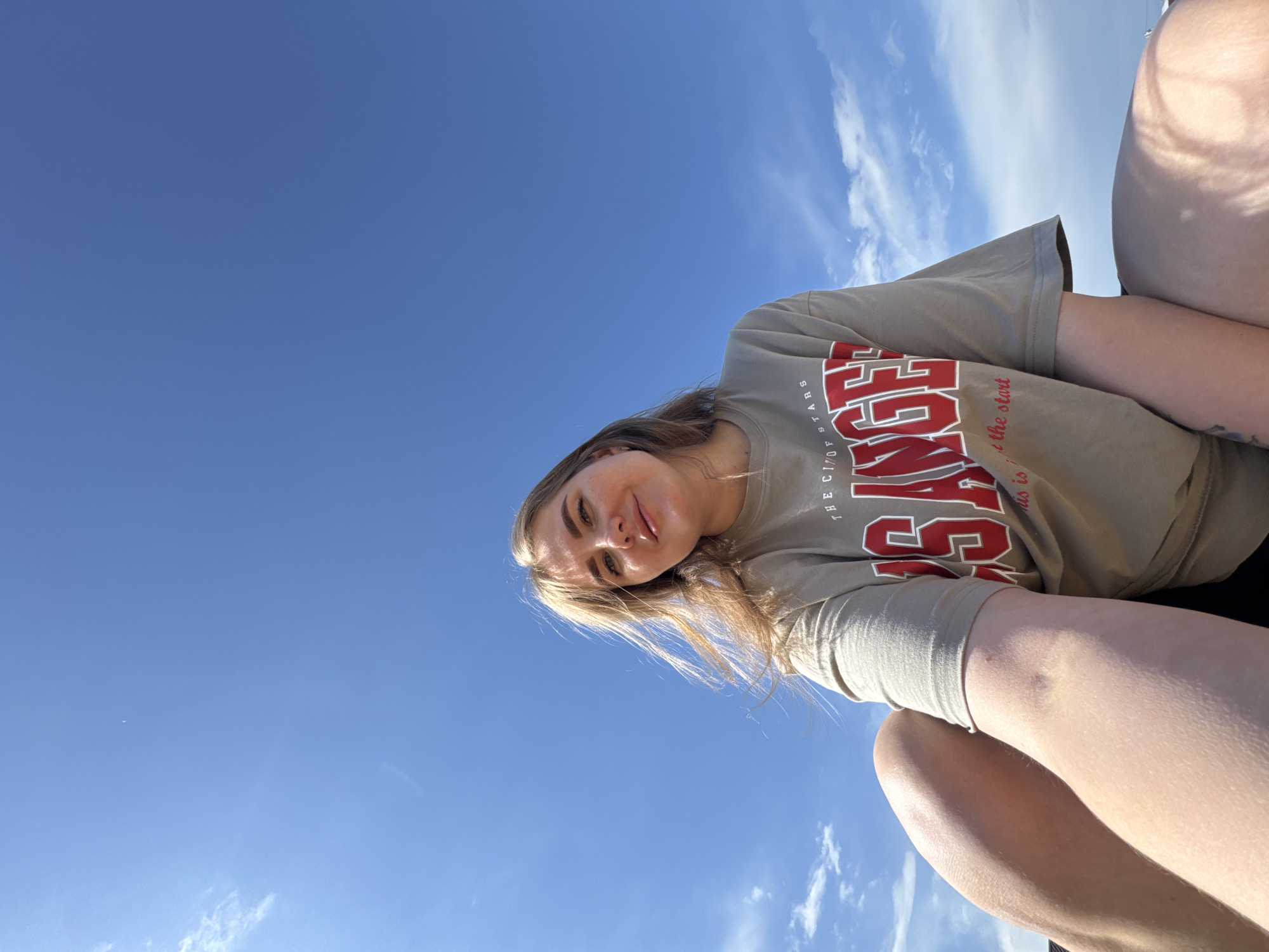 A low-angle shot depicts a young woman against a blue sky with scattered clouds. She is wearing a light-brown t-shirt with the word "ANGELES" in red letters, partially visible. The location is not specified in the city of Ферма, Russia. The woman is angled towards the camera, showing her face. Parts of her legs are visible in the foreground. The lighting indicates a sunny day.