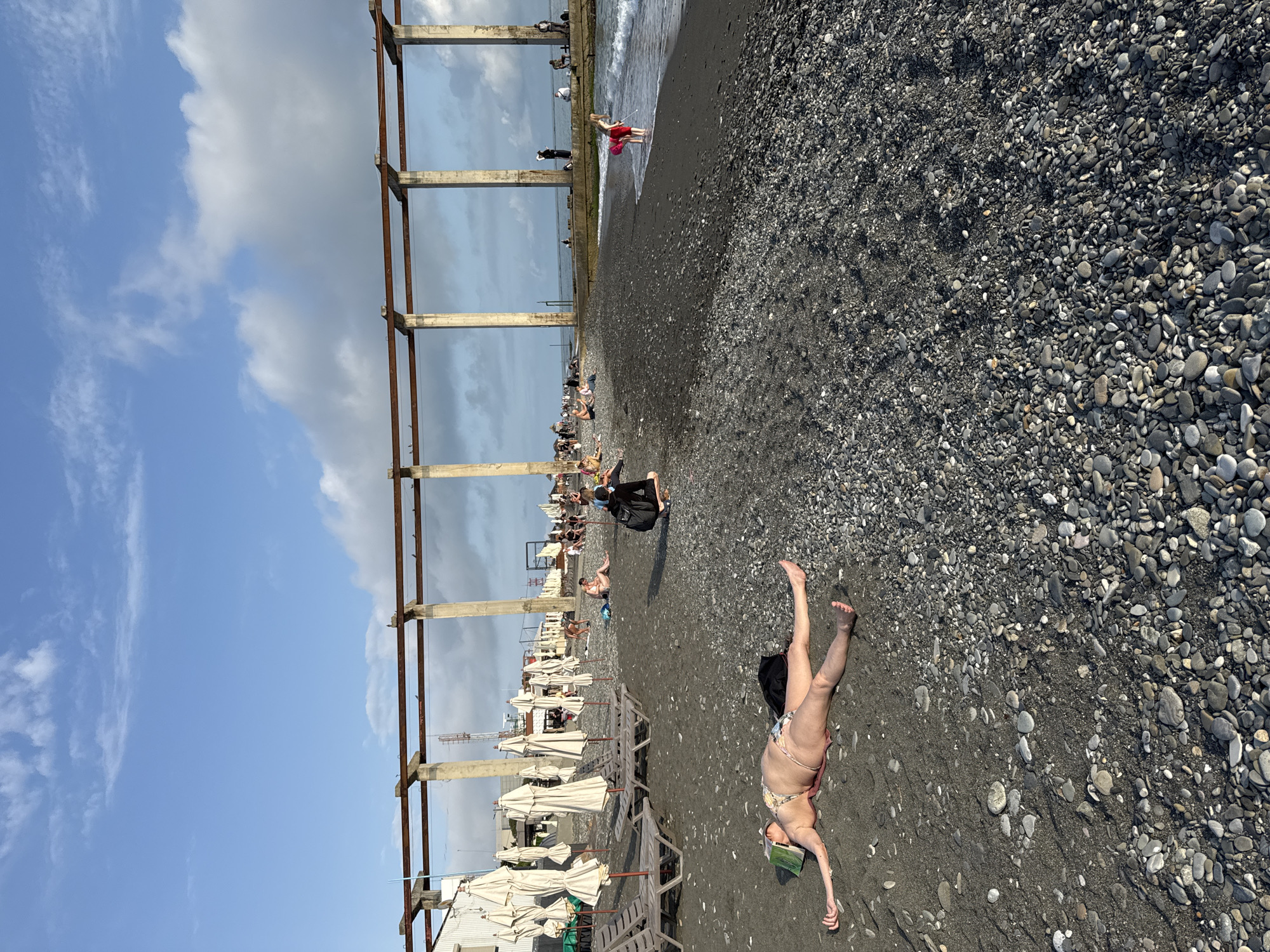 The image depicts a pebble beach in Sochi, Russia. Along the shore, metal infrastructure supports what may have been a pier at one time. People are seen scattered along the beach: some are sitting or lying down, and others are standing near the water's edge. Beach umbrellas and lounge chairs are visible. In the foreground, a woman in a bikini is lying on the beach with a book on her head. The sky alternates between clear blue and cloudy.