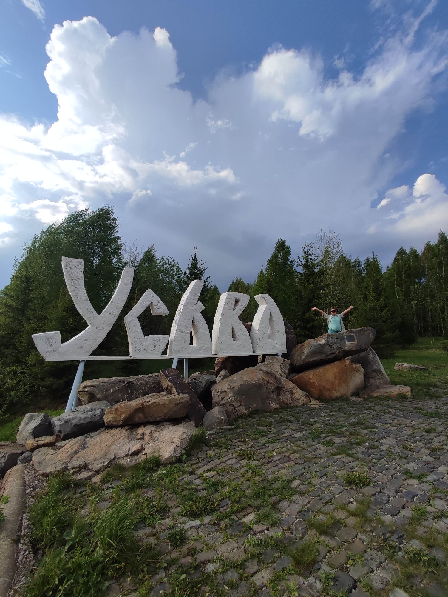 A wide, outdoor shot features a large, white sign spelling out "Усьва" (Usva) in Cyrillic script, located in Gremyachinsk, Russia. The sign is constructed from what appears to be concrete or a similar material, with each letter positioned atop a cluster of large rocks. A woman stands on the rocks next to the final letter, her arms outstretched in a celebratory pose. She is wearing sunglasses and a light-colored outfit.
The sign and rocks are set against a backdrop of lush green trees. Above, the sky is filled with a mix of white and gray clouds. The ground in the foreground is paved with small stones or bricks, and patches of green vegetation are visible.