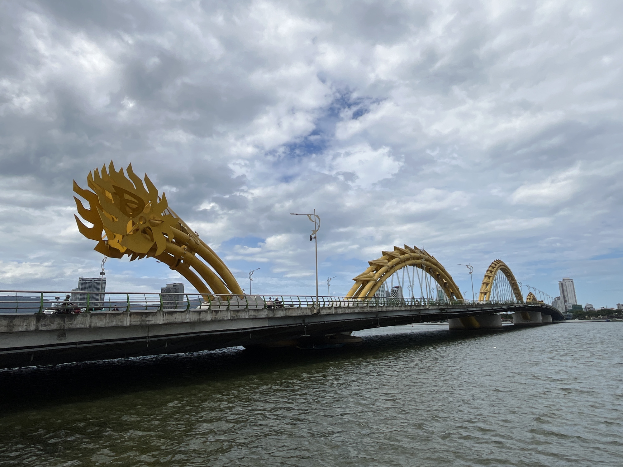 The image displays the iconic Dragon Bridge (Cầu Rồng) in Đà Nẵng, Vietnam, spanning a wide body of water, likely the Han River. The bridge is a prominent golden structure, designed to resemble a dragon, with its head on the left side featuring intricate scales and flame-like elements. Its body forms several archways that support the roadway.
The sky is overcast with a mix of dense grey clouds and some brighter patches, indicating a cloudy daytime setting without direct sunlight. On the bridge's road surface, a few individuals on motorcycles or scooters are visible, engaged in transit across the river. The bridge is equipped with green railings and decorative streetlights.
In the background, modern city buildings, including several tall structures and skyscrapers, are visible on both banks of the river, defining the urban setting. The water in the foreground and beneath the bridge is dark with visible ripples, reflecting the subdued light from the cloudy sky. No discernible text is present in the image.