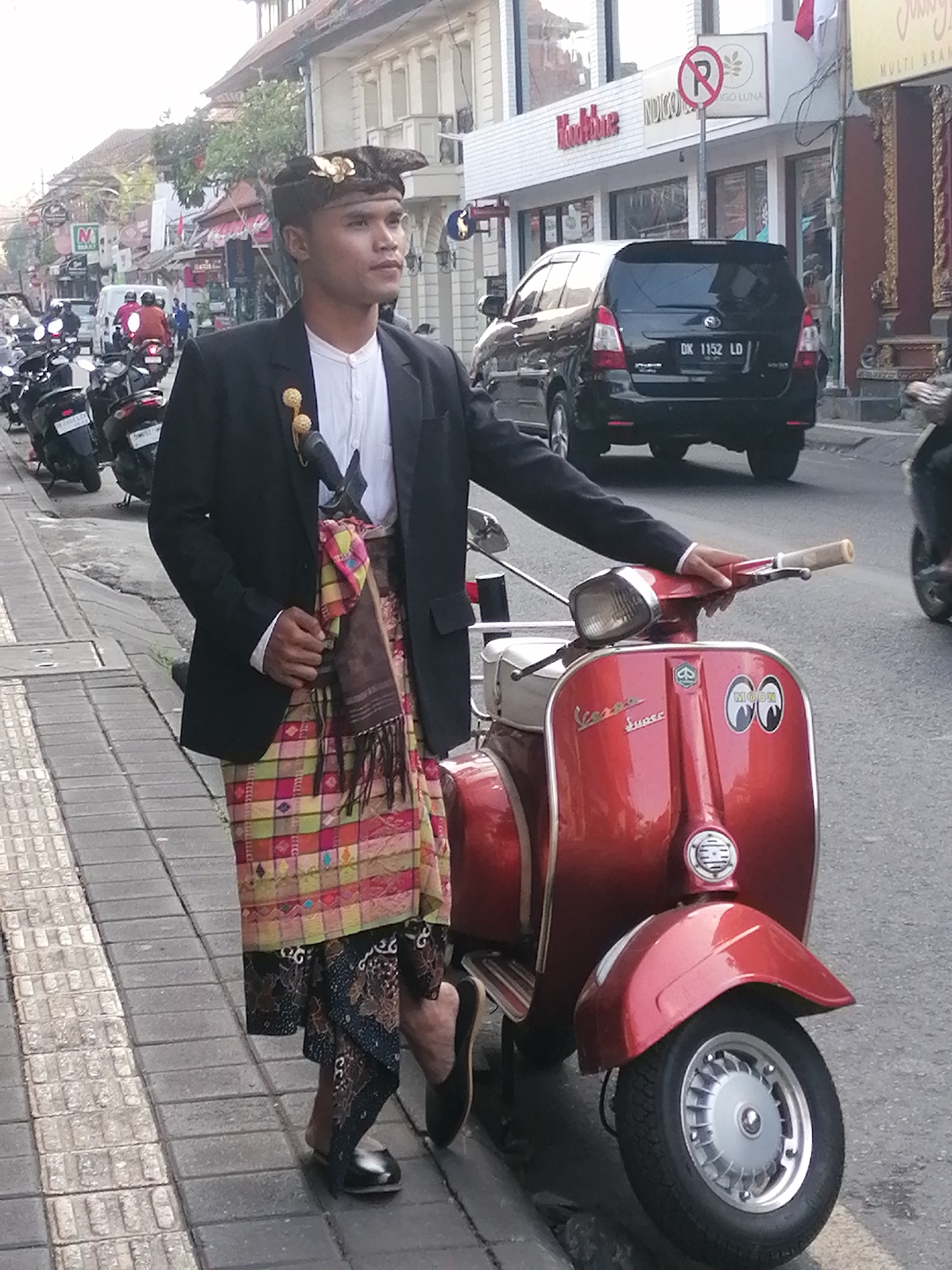 A man wearing a traditional Balinese headdress, black blazer, white shirt, and colorful sarong stands next to a red Vespa scooter on a street in Ubud District, Indonesia. The man has a ceremonial dagger tucked into his sarong. The street is lined with shops and other vehicles, including motorcycles and a black SUV. The man is looking off to the right, and his hand is resting on the scooter's handlebars. The ground is paved with stone tiles, and a sidewalk runs alongside the street. The lighting suggests it is daytime, with shadows cast on the ground.