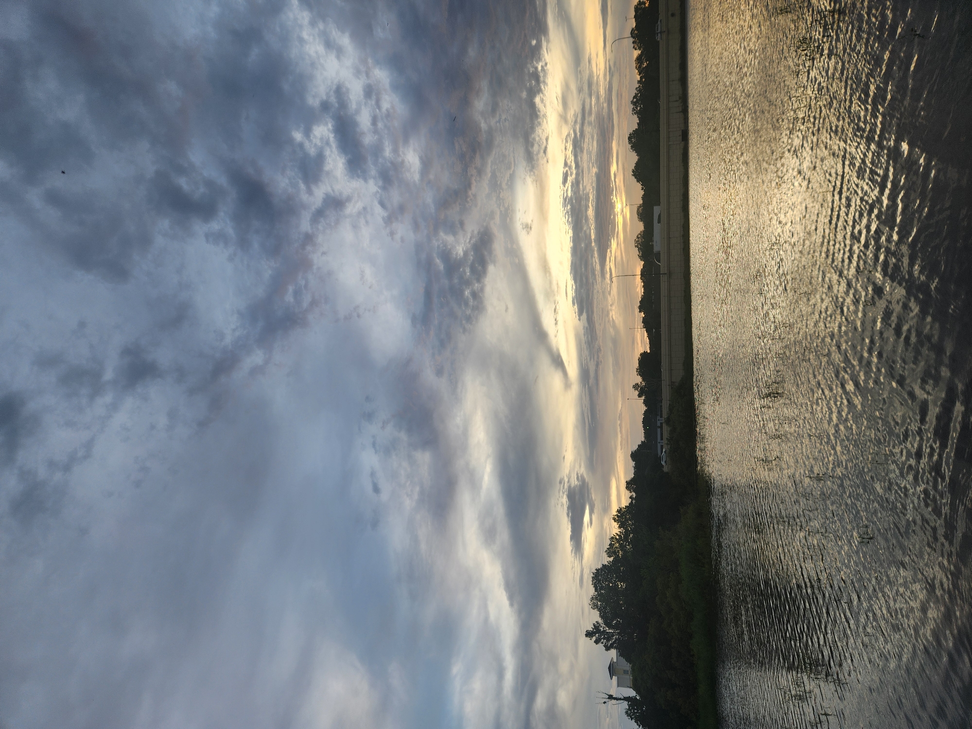 A wide shot captures a body of water with rippling reflections of the sky. The sky is filled with dramatic clouds, with a sliver of yellow light peeking through near the horizon, suggesting either sunrise or sunset. Along the far bank of the water, a dense line of trees is visible, with a modern building or structure nestled among them on the left. Beyond the trees, a highway or elevated roadway runs horizontally across the frame, with traffic lights and a few vehicles discernible. The overall scene appears to be a natural landscape within an urban or suburban setting, possibly a retention pond or lake near a developed area. The image was taken in Altamonte Springs, United States of America.