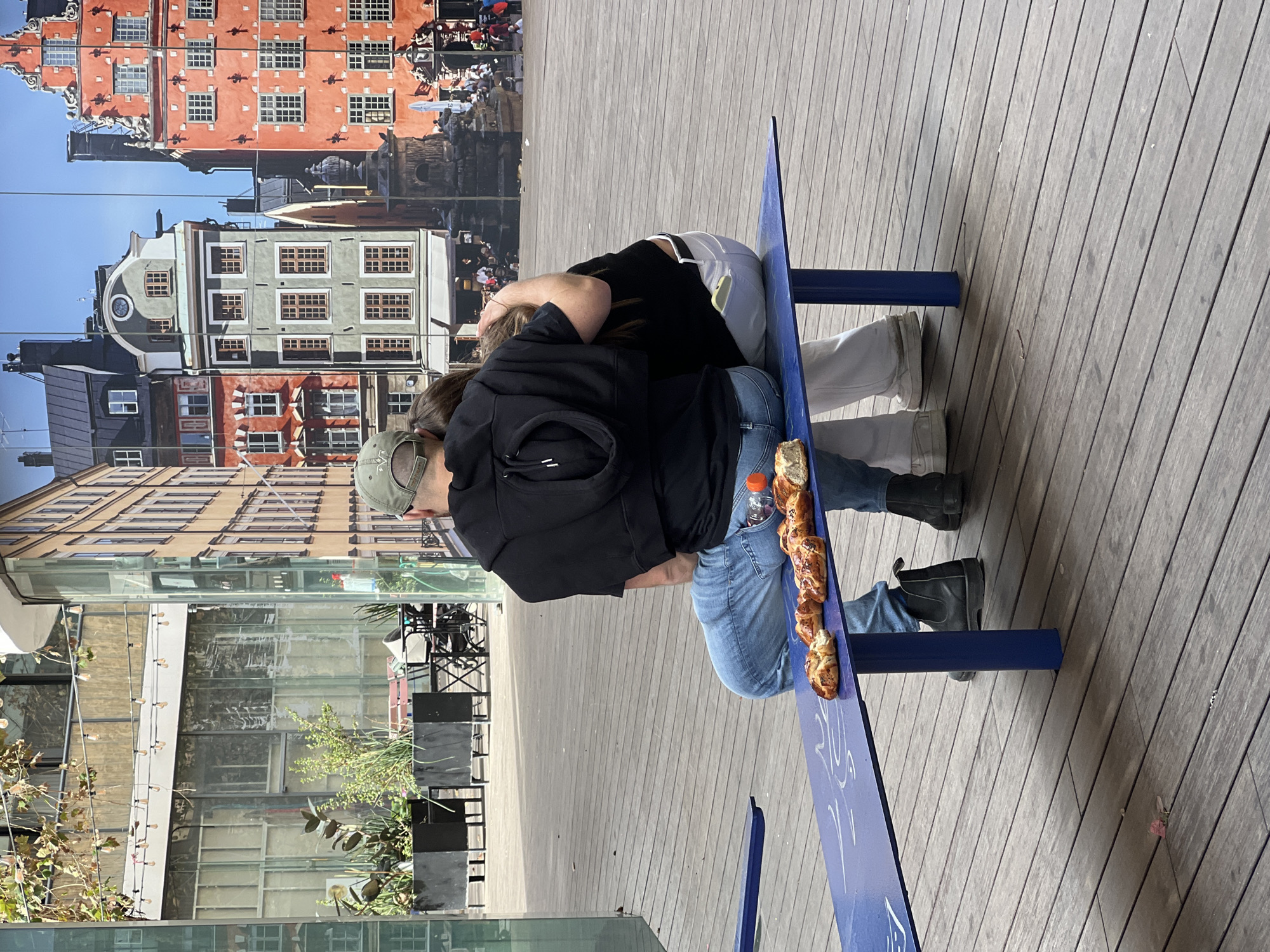 An overhead outdoor shot depicts two individuals seated on a blue metal bench in the Sarona Market complex in Tel-Aviv, Israel. The ground is covered with wooden decking. Four baked goods, resembling pastries or sweet buns, are placed on the bench in front of the individuals, along with a clear plastic water bottle.
The male, on the left, wears a green baseball cap, a black hooded sweatshirt, blue jeans, and black boots. His left arm is extended around the shoulders of the female. A light-colored object is partially visible in his right rear jeans pocket.
The female, on the right, wears a black top, light-colored pants, and white sneakers. Her head is resting against the male's shoulder. A yellow-cased mobile phone is visible in her left rear pants pocket.
In the mid-ground to the left, black planter boxes containing green foliage are visible, along with blurred elements of an outdoor café area including tables and chairs. String lights are suspended overhead. The background is dominated by a large, reflective glass surface that displays a vibrant reflection of a European-style street scene featuring multi-story buildings in red, grey, and brown hues, characterized by numerous windows and architectural details. A clock is visible on one building, and an ornate, dark monument or statue is also part of the reflection, suggesting a public square with distant, indistinct figures.