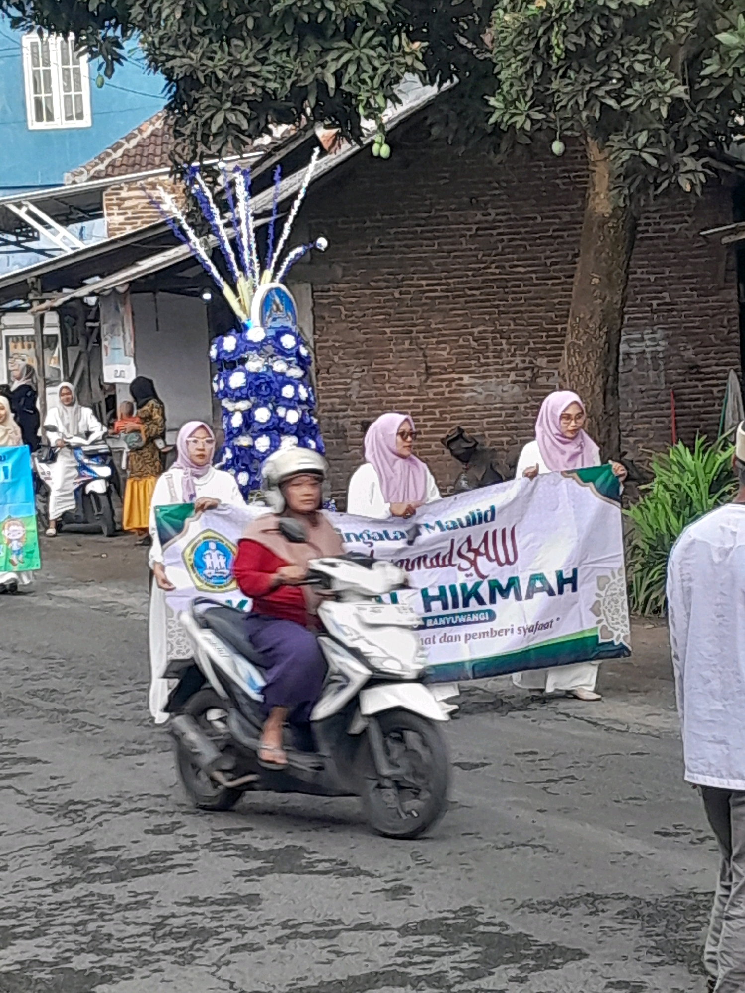In Gentengwetan, Indonesia, a parade is underway on a paved road. A group of women, most wearing hijabs, are participating, some on motorcycles and others walking while holding banners. One woman rides a white scooter, wearing a red top and purple trousers, with a helmet. Another woman in the foreground holds a banner that reads "Maulid Muhammad SAW HIKMAH BANYUWANGI". Behind them, a tall, blue and white floral decoration is visible, possibly part of the parade's float. The buildings in the background are made of brick, and a tree with hanging fruit is present. The scene captures a cultural or religious event in the town.