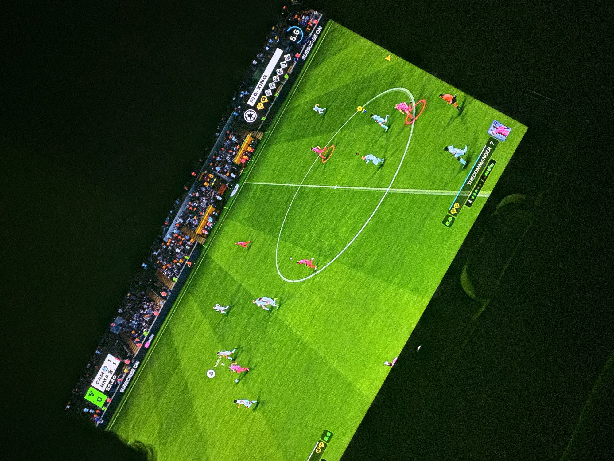The image shows a screen displaying a soccer video game. The game is in progress, with teams identified as CAM and BMA with a score of 1-1 at 32:20. The game features virtual players on a green field with white lines and a center circle. The player avatars are depicted in light blue and pink uniforms. Along the left side of the screen, there is a virtual crowd. The user interface displays information such as the player name "THECOMMANDER 7," ratings (8.0), and other game stats. The text "SUBSCRIBE ON Youtube" appears twice. The name "99 G. XING" is also present. The setting appears to be indoors with low ambient light. No specific location within Welshpool is identifiable from the image.