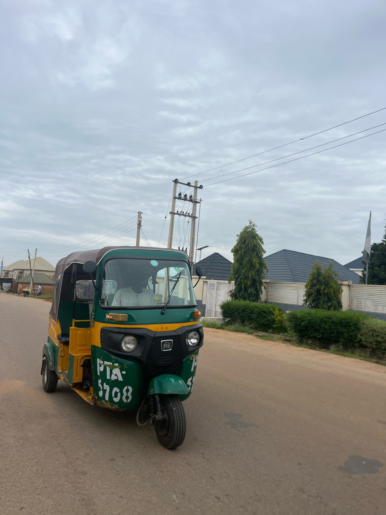 A yellow and green three-wheeled auto-rickshaw, identified by the license plate "PTA-5708," is parked on an asphalt road in Barakin Akawo, Nigeria. The auto-rickshaw is positioned on the right side of the road, facing forward. A person wearing a light-colored shirt and a face mask is visible in the driver's seat. The vehicle has a dark-colored canopy. Behind the auto-rickshaw, a street with several residential buildings and lush green trees can be seen. Overhead, power lines and utility poles are present, suggesting an urban or suburban environment. The sky is overcast with a diffused light. The overall scene depicts a typical street view in a Nigerian town.