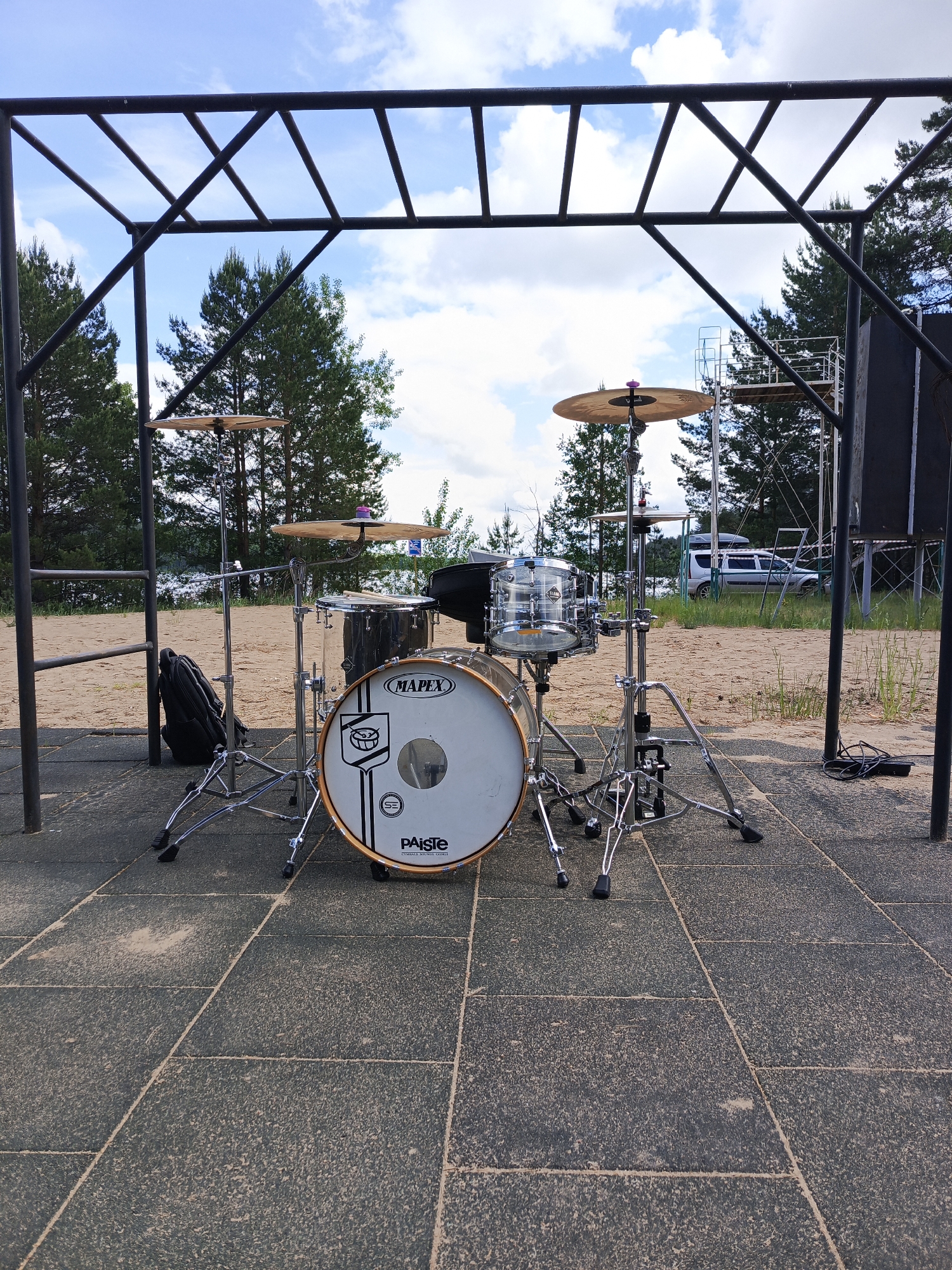 The image shows a drum set placed outdoors on a paved area. The drum set includes a bass drum, snare drum, and cymbals. A black backpack is positioned to the left of the drum set. Above the drums, a metal frame structure is visible. The background includes trees, a body of water, and a glimpse of a white van. The setting appears to be a public space with outdoor fitness equipment.