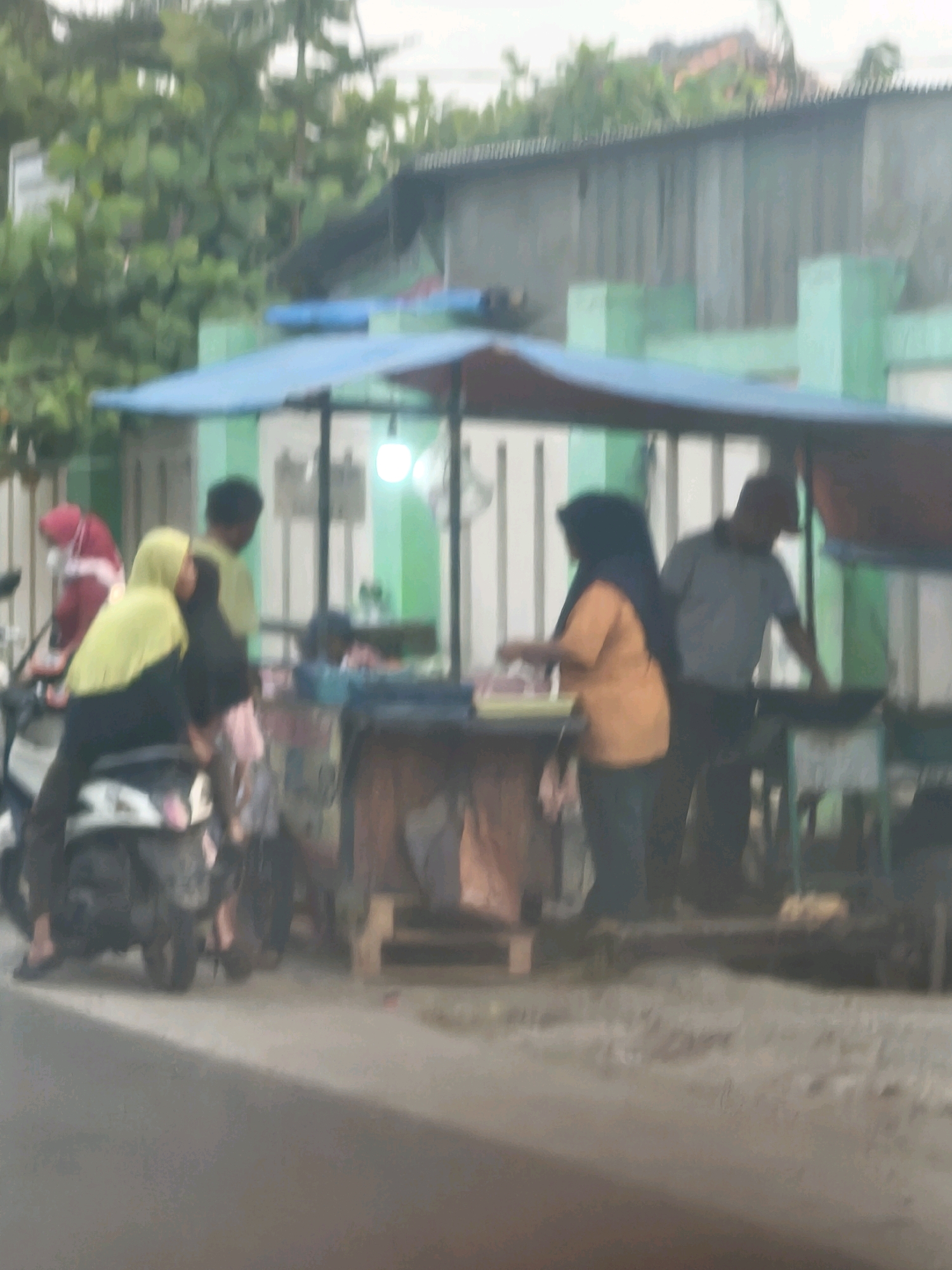 A slightly blurry, medium-distance shot captures an outdoor scene on a street in Satriamekar, Indonesia. A food stall with a blue tarp roof and green vertical supports is set up along the side of the road. Several people are visible around the stall.
In the foreground, on the left, two individuals are seated on a motorcycle. One person, wearing a bright yellow headscarf and dark clothing, is leaning forward. Another person, partially obscured, wears a red headscarf and dark clothing.
Behind the motorcycle, a person in a light green shirt stands near the stall. To the right of the stall, a woman in an orange shirt and dark headscarf is engaged in an activity, possibly preparing food. Next to her, a man in a grey t-shirt stands, looking down towards something on the stall.
The stall itself has a wooden counter displaying various items, including what appear to be food containers. The environment suggests a typical street scene in Indonesia, with lush green foliage visible in the background and a wall behind the stall. The ground is paved, and the overall lighting is overcast.