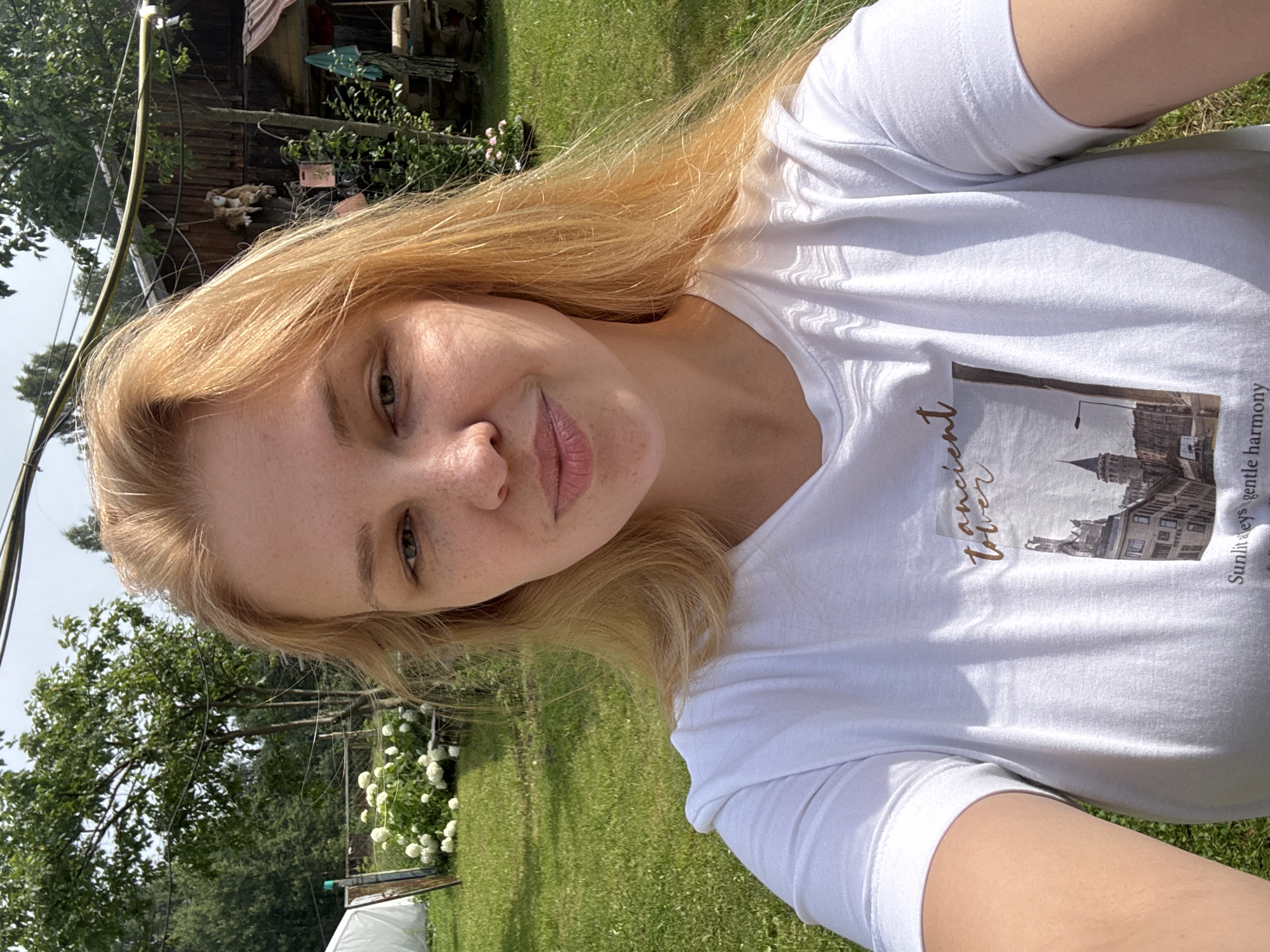 A blonde woman with light skin and freckles smiles at the camera in a selfie. She is wearing a white t-shirt with a graphic design featuring a building and text. The background appears to be a rural or suburban backyard with green grass, trees, and a wooden structure that resembles a shed or small building. There is also a large bush with white flowers to the left of the frame. The overall lighting suggests it is daytime and sunny. The location is not discernable from the image itself.