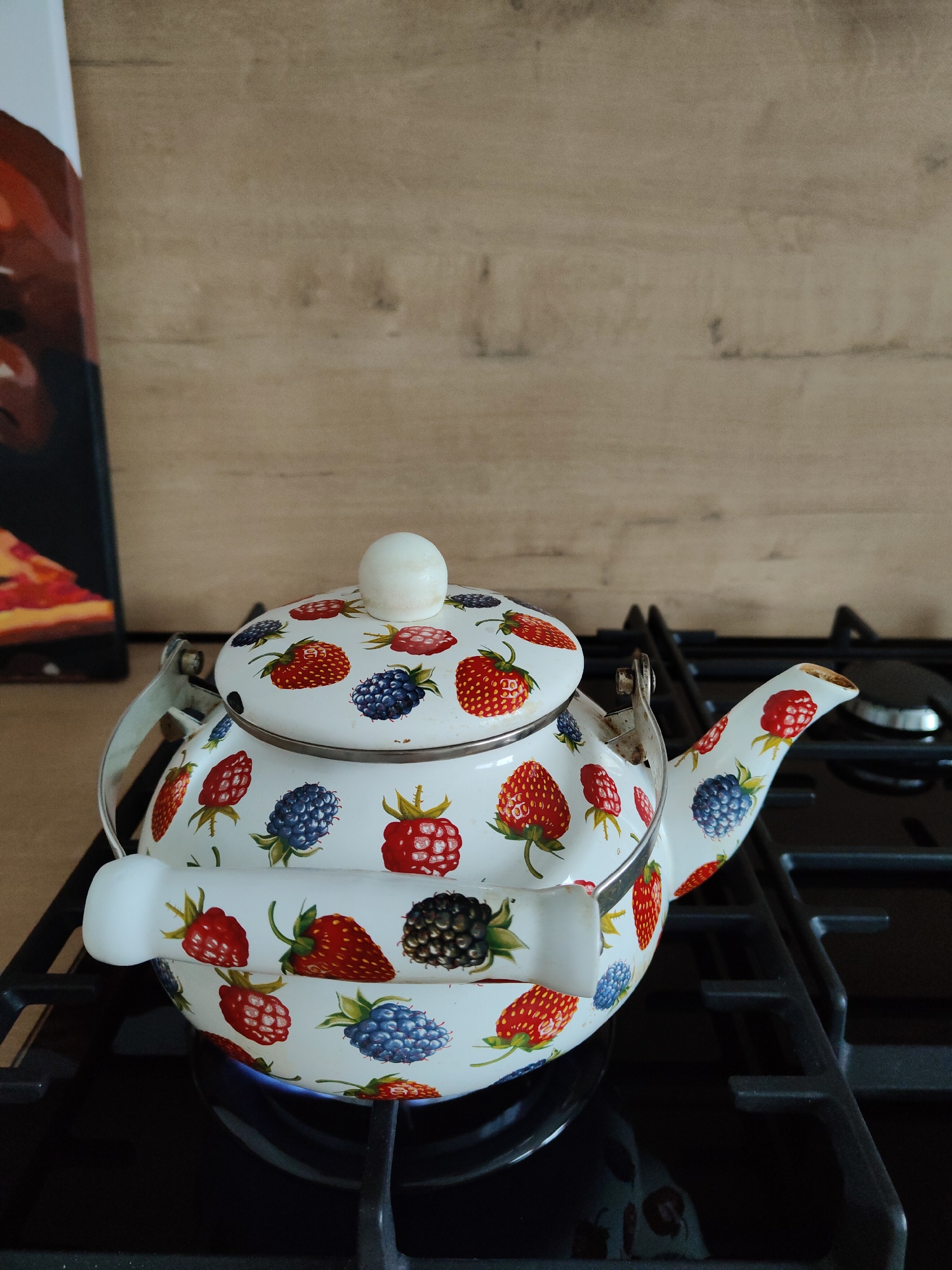 A white teapot decorated with illustrations of strawberries and blackberries sits on a black gas stovetop. The burner beneath the teapot is lit, producing a faint blue flame. The teapot has a white lid with a rounded knob and a white handle. In the background, there is a wooden wall and part of a framed picture, possibly a painting, is visible. The location of the scene is Unknown, Unknown.