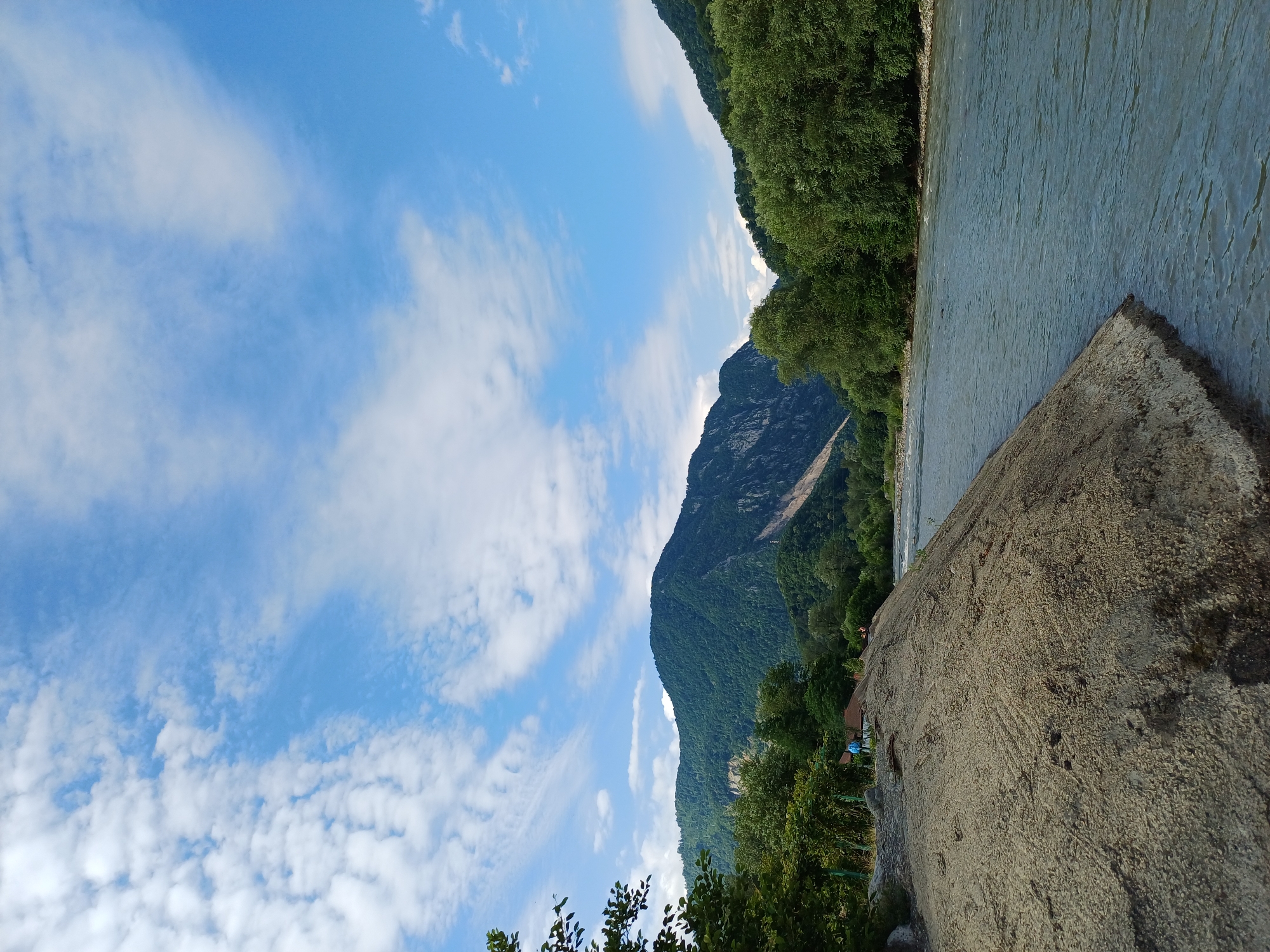 A wide shot captures a natural landscape under a bright blue sky dotted with white clouds. In the foreground, a large mound of coarse, light-colored sand dominates the left side, with a textured surface showing small pebbles and some darker, wet patches towards the bottom. To the right of the sand mound, a wide, flowing river with a choppy surface stretches into the distance, its water a muted blue-gray.
Beyond the river, a dense forest lines the banks, with lush green trees forming a thick canopy. In the mid-ground, a large, imposing mountain rises, its slopes covered in dark green vegetation. The mountain has a jagged, rocky face on its right side, suggesting a steep incline or cliff. Further back, more mountains and hills are visible, also covered in trees. The sky above is clear blue with scattered white, wispy clouds, particularly concentrated on the left side.
In the distance, near the riverbank and nestled amongst trees, a few small structures, possibly buildings, are visible. A few people can be seen as small figures, including one person wearing a blue backpack standing near the structures on the left bank. Another person is visible further along the bank. The scene suggests a peaceful, natural setting, likely a popular spot for outdoor recreation given the presence of people and the sandy area that could serve as a resting or gathering place. The image appears to be taken on a sunny day.