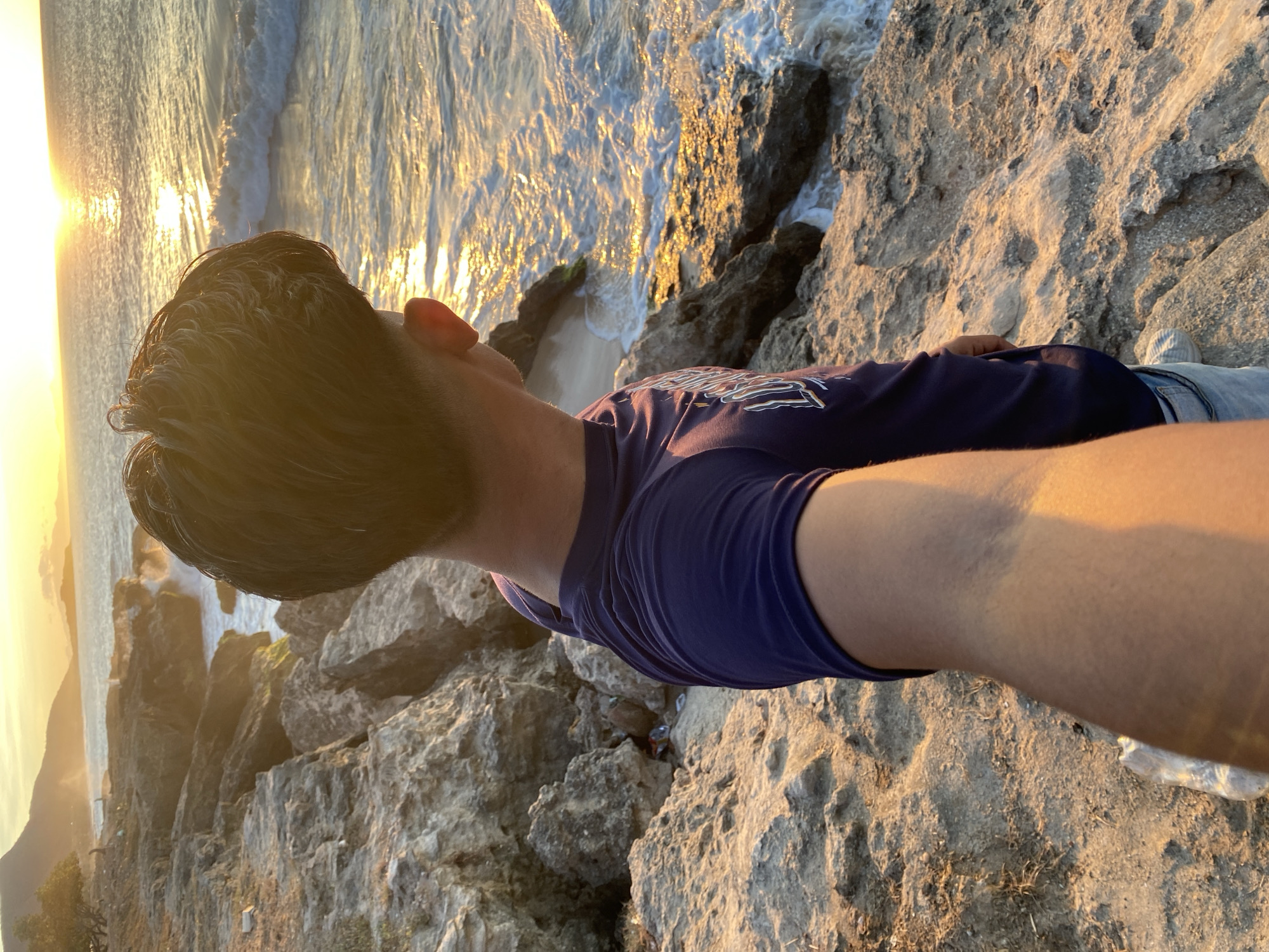 A person is lying on a rocky outcrop overlooking the ocean in what appears to be a coastal setting. The person is wearing a dark t-shirt and light-colored jeans. The rocks are light-colored and weathered, with the ocean visible in the background. The sun is setting, casting a warm glow over the scene. Based on the location provided, this is likely in or around Juan Griego, Venezuela, due to the typical characteristics of its coastal landscape. The person is likely taking a selfie or otherwise documenting the view.