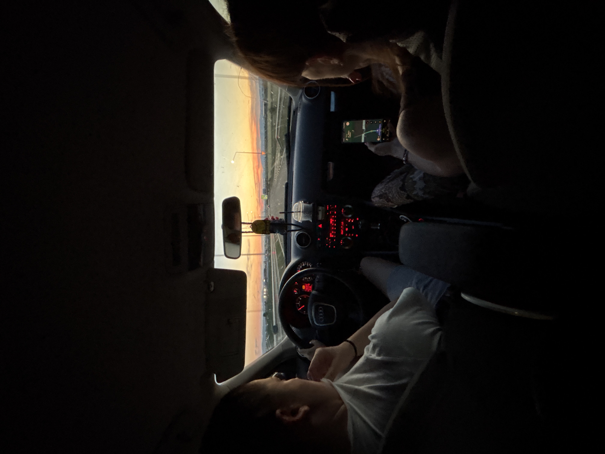 In Sofia, Bulgaria, at dusk, two individuals are inside a car. The person on the left is driving, and the person on the right is looking at a smartphone. Both individuals' faces are partially obscured, but the scene suggests a journey along a highway with a sunset visible in the distance. The car's dashboard displays illuminated red lights, and the interior is dimly lit.