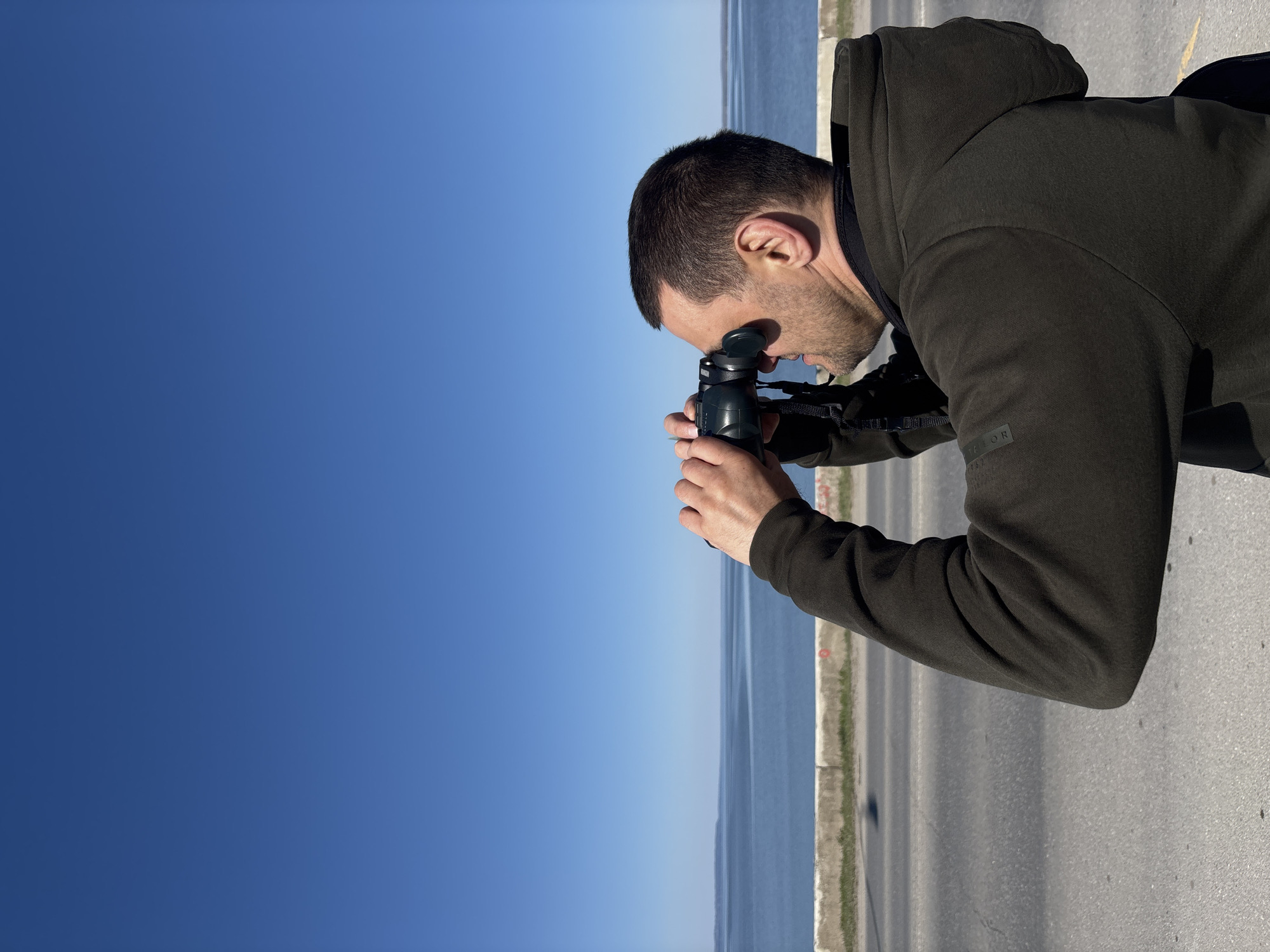 A man is using binoculars on a sunny day. He is wearing a dark green hoodie and standing on an asphalt road. In the background, there is a body of water and a clear blue sky. The man's location is in the city of Chaikovsky, Russia.