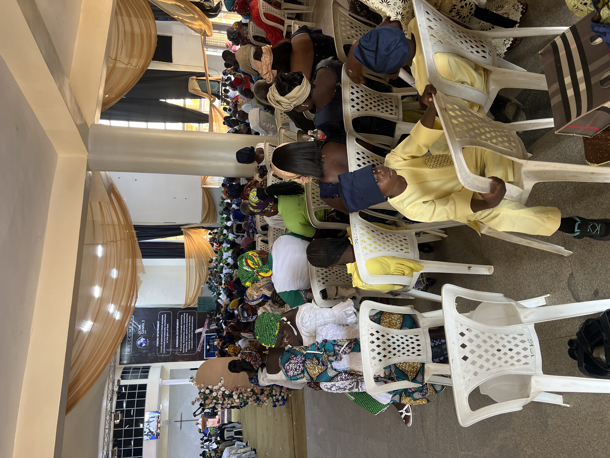 The image depicts an indoor gathering, likely a religious service, taking place in a hall in Kabong, Nigeria. A large congregation of people, predominantly dressed in vibrant attire and headwraps, are seated on rows of white plastic chairs. The hall is decorated with draped fabric in shades of gold and dark grey, and banners with text such as "WELCOME TO ECWA GOSPEL 2" and "VISION STATEMENT" are visible on the wall. In the foreground, two young boys in yellow outfits and blue head coverings are seated. The overall atmosphere appears to be one of communal assembly.