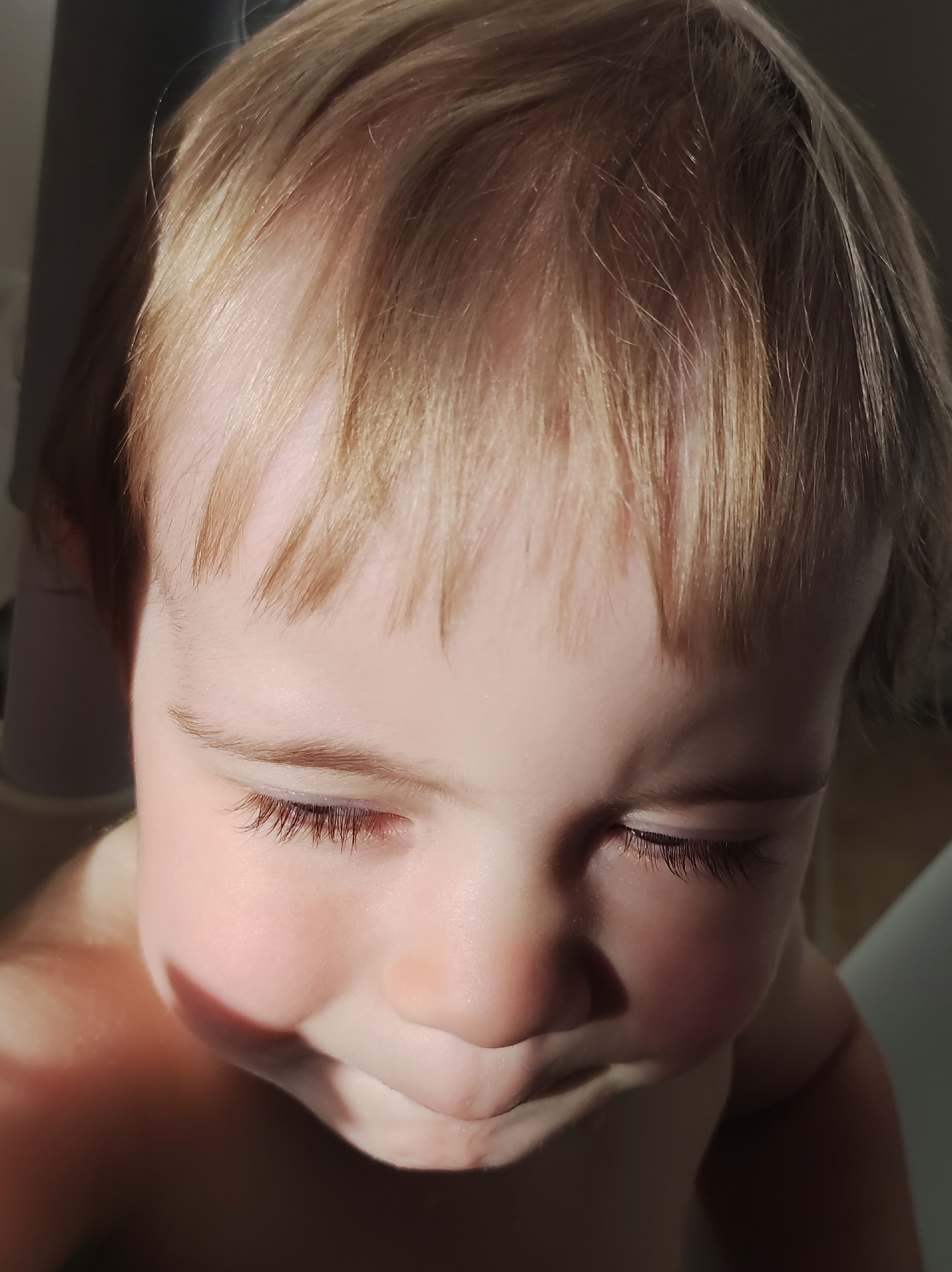A close-up shot features a young child with fair hair and closed eyes. The child's face is well-lit on one side, casting shadows on the other. The child appears to be fair-skinned with long, dark eyelashes. The image captures a tender moment, focusing on the child's peaceful expression. The location is unknown and not indicated in the picture.