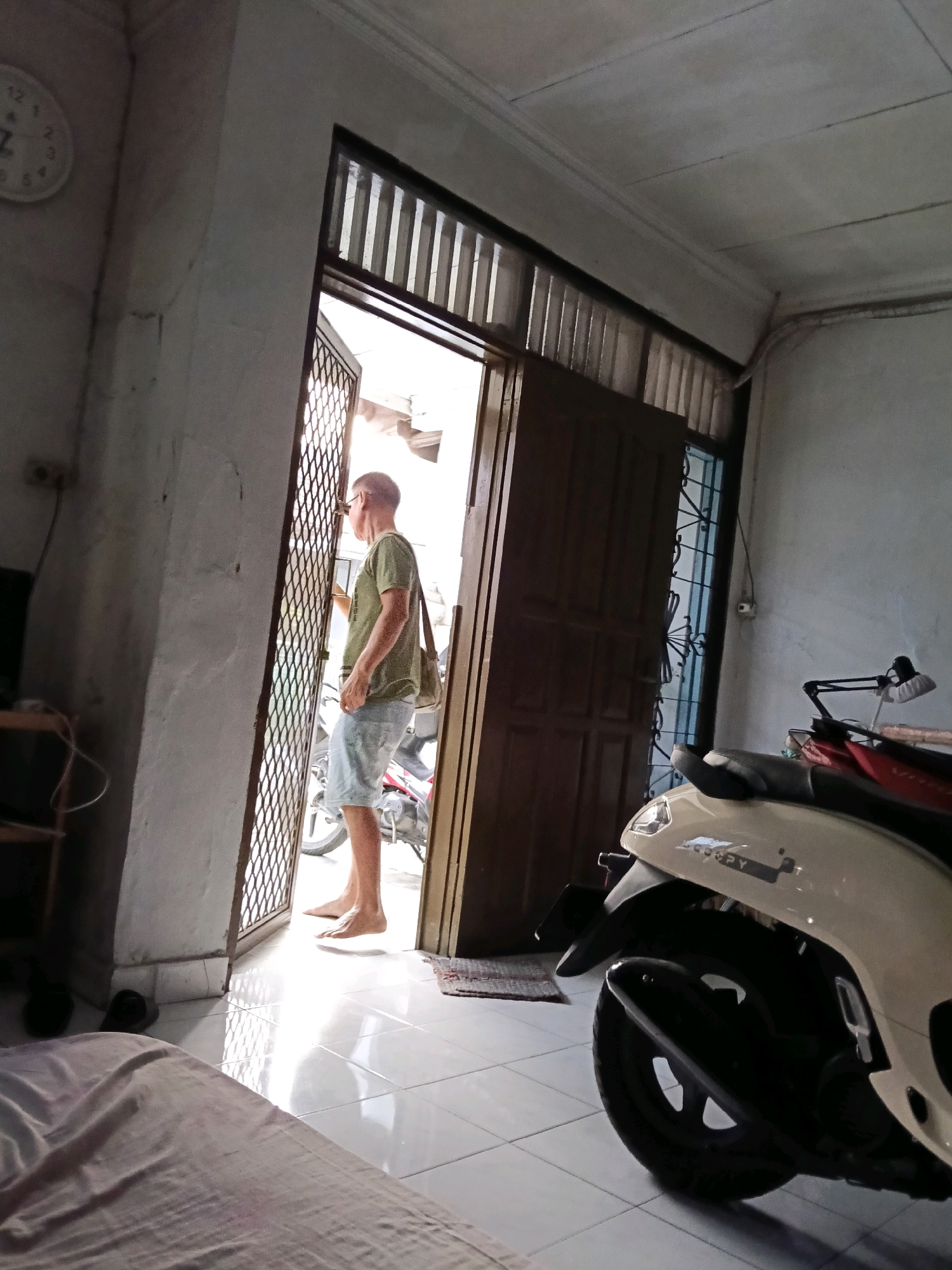 The image depicts an indoor scene, likely a residential dwelling, with a man standing in a doorway that leads outside. The man, viewed from the side, wears a green t-shirt and light blue denim shorts. He has a bag slung over his shoulder. His feet are bare. Outside the doorway, a motorcycle is partially visible.
The interior features white tiled flooring, and a section of a bed or mattress covered in a light pink fabric is in the foreground on the left. A clock is mounted on the wall to the left of the doorway. The walls appear to be painted white or a light color, and the ceiling is also white. The doorway itself is made of dark wood and has a grate-like screen.
The overall lighting suggests it is daytime, with natural light coming through the open doorway. The environment suggests a simple, functional living space. The location is identified as Jatimulya, Indonesia.