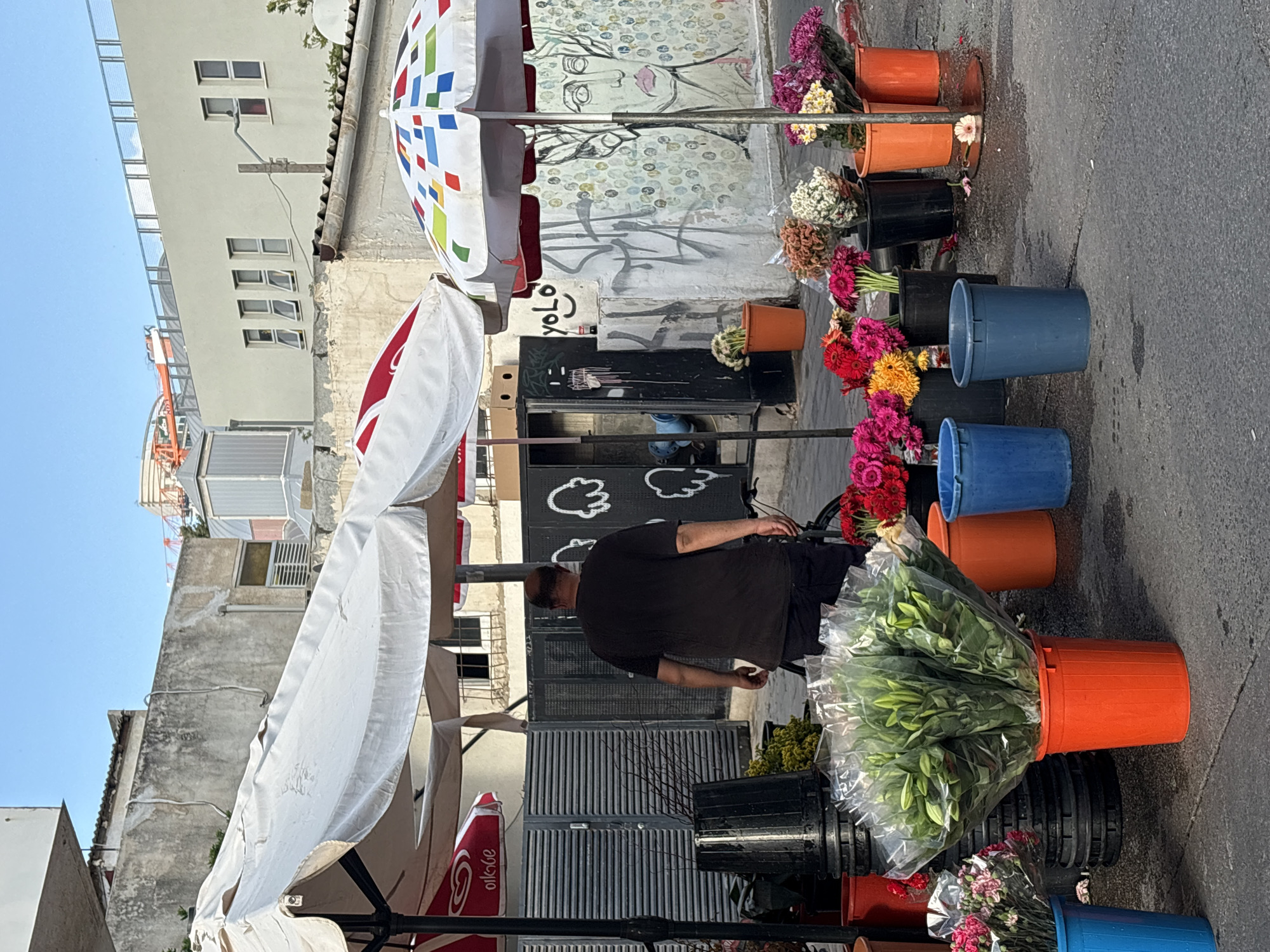 The image depicts a man standing at a flower stand located on a street in Tel Aviv, Israel. The man is wearing a black t-shirt and shorts. He appears to be arranging or tending to the flowers. The flower stand is comprised of multiple buckets filled with various types of flowers in an array of colors. The buckets are arranged in a vertical display, suggesting a small, pop-up style flower shop. Behind the flower stand, there is a wall with a faded mural and graffiti. Several umbrellas are providing shade for the flower display. The background buildings are typical of Tel Aviv architecture.